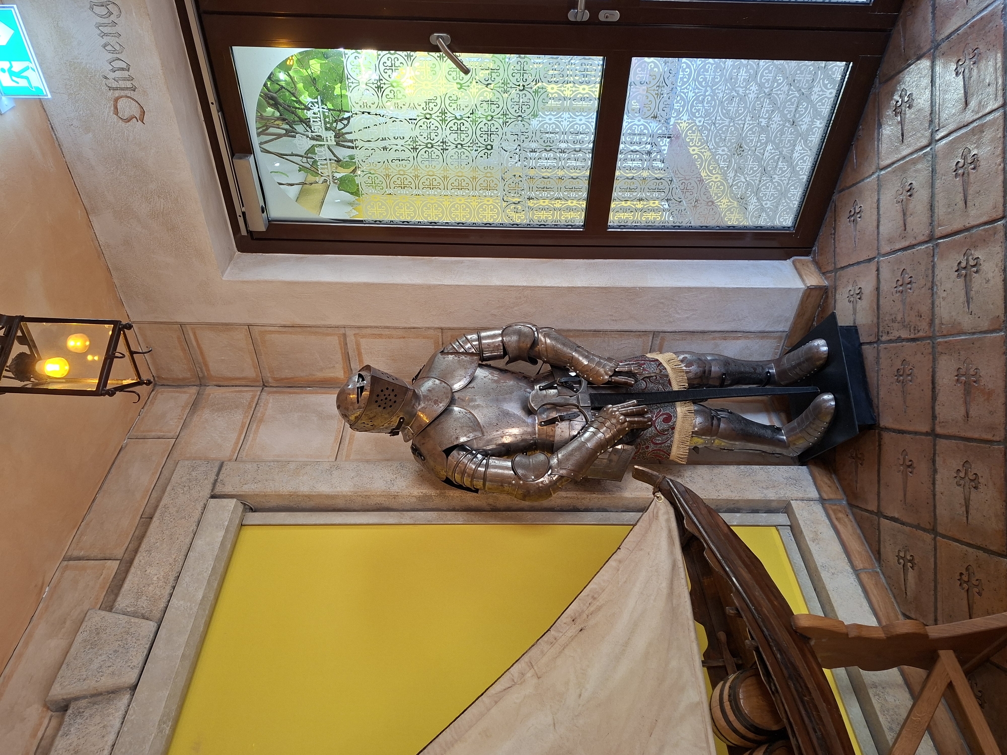 The image depicts a corner of an indoor space, possibly a restaurant or a historical-themed establishment in Rheinhausen, Germany. The scene is dominated by a full suit of armor standing to the right of a bright yellow, recessed doorway or feature. The armor is made of metal, polished to a shine, and stands upright on a black pedestal. It is adorned with a decorative, fringed fabric belt around the waist.
To the left of the armor, a wooden model boat with a white sail is displayed. The boat rests on a wooden stand and contains two small barrels. The wall behind the boat is bright yellow.
The room's architecture features stone-like textured walls in neutral tones and a tiled floor with a decorative pattern. To the right of the suit of armor is a large glass door with a dark brown frame. The glass is partially obscured by a decorative etched pattern featuring repeating circular motifs. Above the door, an illuminated green emergency exit sign with a running figure is visible. Further up on the wall, the word "Oliveng..." is faintly inscribed in a decorative font.
The lighting in the scene appears to be a mix of ambient room light and light from a decorative pendant lamp with three bulbs hanging from the ceiling in the upper center of the image. The overall impression is one of a curated display designed to evoke a historical or themed atmosphere.