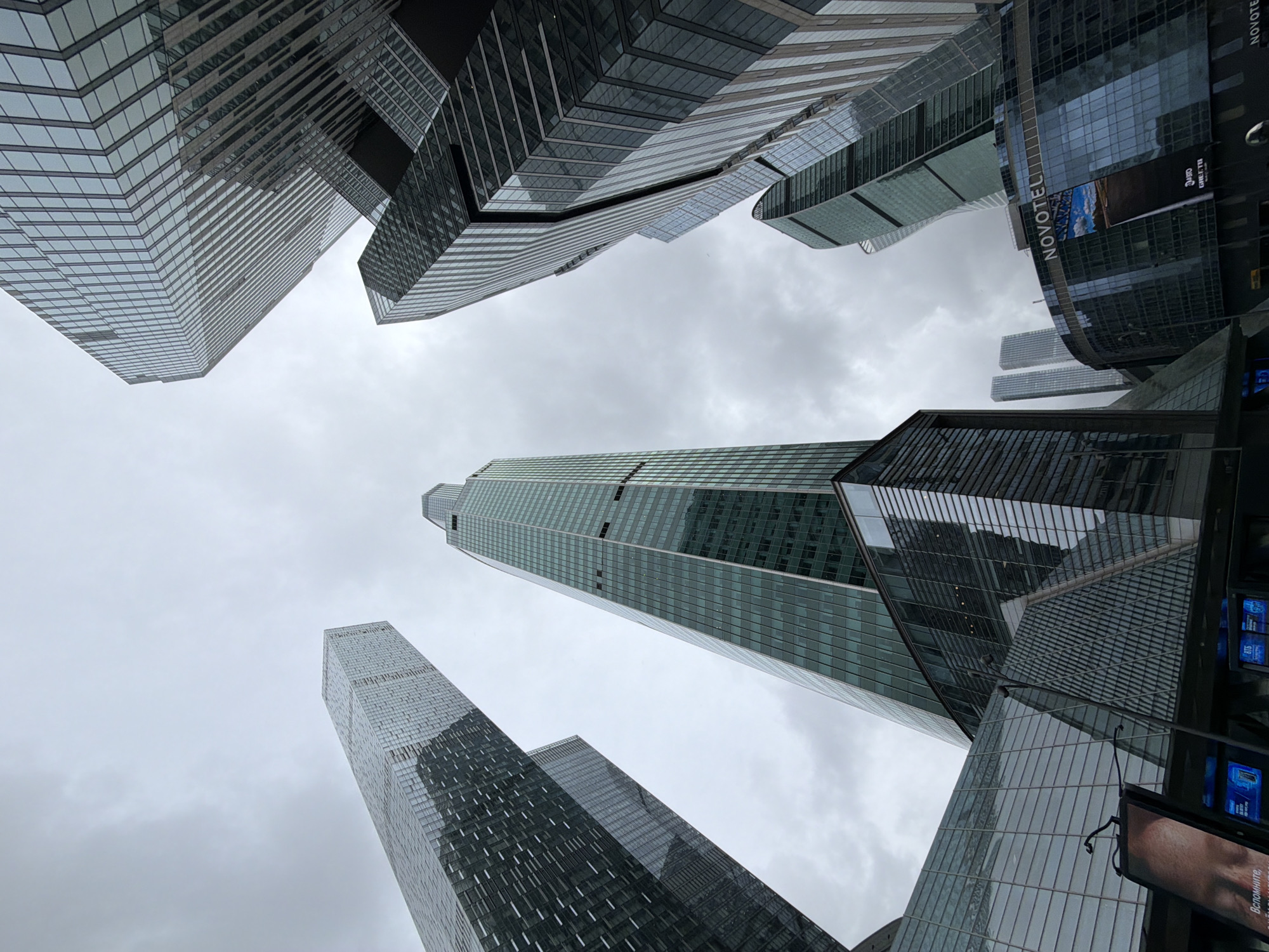 The image presents a dramatic upward view of several modern skyscrapers against an overcast sky, characteristic of a contemporary urban landscape. The perspective emphasizes the towering height and angular geometry of the buildings.
The primary subjects are multiple high-rise structures, predominantly clad in glass and metal. These buildings display diverse architectural forms, including sharply angled facades, stepped designs, and sleek, continuous surfaces that reflect the muted light of the cloudy sky. A prominent, slender skyscraper occupies the central-right portion of the frame, featuring extensive glass paneling. To its left, another building shows a complex, faceted design with numerous horizontal lines.
On the right side, a distinctive curved building is visible, prominently featuring the white "NOVOTEL" logo. Below this, a large digital display shows partial text including "3PRO GILLETTE" and a fragment of Cyrillic text that appears to start with "Вспомните" (meaning "Remember"). A smaller digital screen nearby displays "ВТБ", likely referencing VTB Bank. These signs suggest a commercial and business district.
The sky is entirely cloudy and grey, indicating an overcast daytime condition without direct sunlight. There are no people or specific activities depicted, lending a static and imposing quality to the architectural scene. The overall impression is one of advanced urban development and grand scale, consistent with a modern metropolitan area like Khimki, Russia.