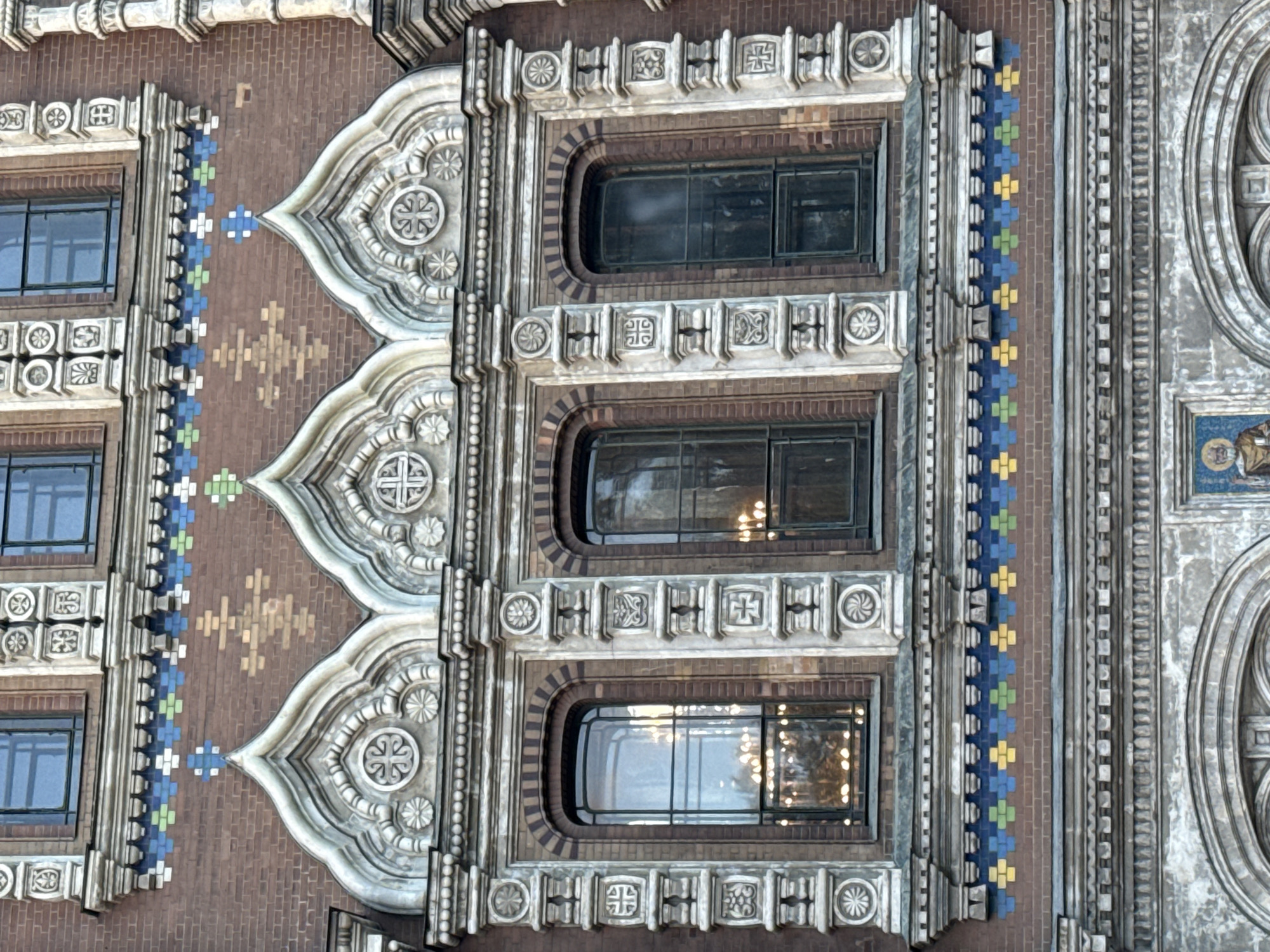 The image shows a detail of the facade of the Church of the Savior on Spilled Blood in Saint Petersburg, Russia. The photograph focuses on a section of the exterior featuring three arched windows set within ornate stone surrounds. The windows are tall and narrow, with dark frames. The brickwork of the building is visible, decorated with bands of colorful mosaic tiles in blue, green, and yellow. Above the windows, the facade is further embellished with decorative stonework, including carved motifs and finials. The reflections in the windows suggest an interior with lighting fixtures. The overall impression is one of intricate architectural detail and vibrant ornamentation characteristic of Russian Revival style.