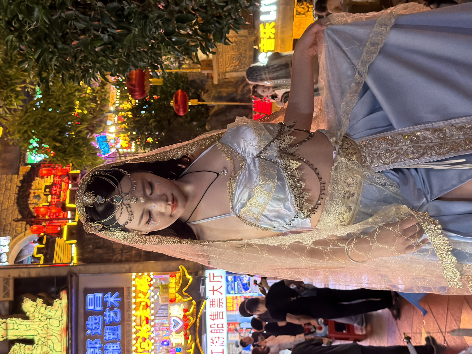 A young woman, adorned in a light blue and gold costume with a sheer veil, stands outdoors at night in a busy street market in Yuzhong District, China. The woman's face is lightly made up with glitter on her eyelids. Her costume includes a decorative headdress, a bejeweled halter top, and a flowing skirt. She is holding the skirt with her right hand. In the background, neon signs illuminate the bustling street, with trees adorned with colorful lights and red lanterns hanging overhead. Several people are visible in the background, some walking and others observing. The scene suggests a festive or cultural event is taking place.