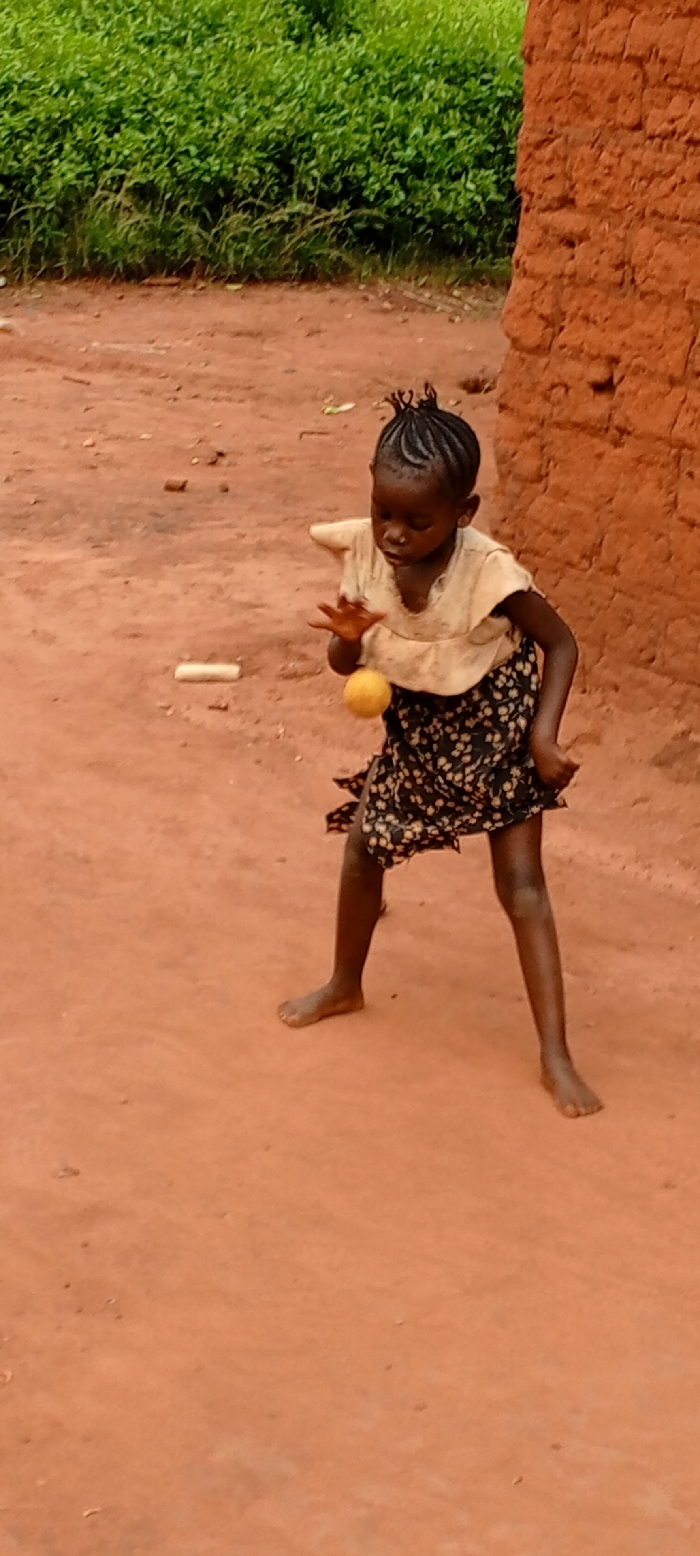 A young, dark-skinned child with braided hair stands barefoot on reddish-brown earth. The child is dressed in a light-colored, short-sleeved top and a dark, patterned skirt. Their legs are spread with bent knees, indicating a dynamic posture. The child's right hand is open with splayed fingers, positioned above a yellow, spherical object, suggesting an action involving the object. A light-colored, elongated object lies on the ground to the child's left. The background features dense green vegetation on the left and a reddish-brown, unplastered brick or mud-brick wall on the right. The scene is located in Ngalula, Democratic Republic of the Congo.