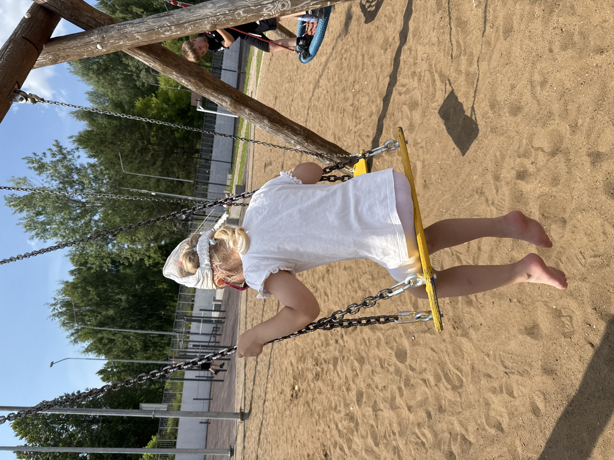 In Chaikovsky, Russia, a young girl with blonde hair, wearing a white dress and a light-colored headscarf, is seated on a yellow swing. She is viewed from behind, with her bare legs dangling towards the sandy ground. To the right, a boy in a black t-shirt and shorts is on a blue rope swing, his back mostly to the camera. The playground features a wooden swing set structure, with metal chains supporting the swings. The environment is characterized by a sandy surface and a background of green trees and a blue sky with scattered clouds. In the distance, there are metal bars and structures, likely part of an outdoor exercise area, and some fencing. The scene appears to be set on a sunny day.