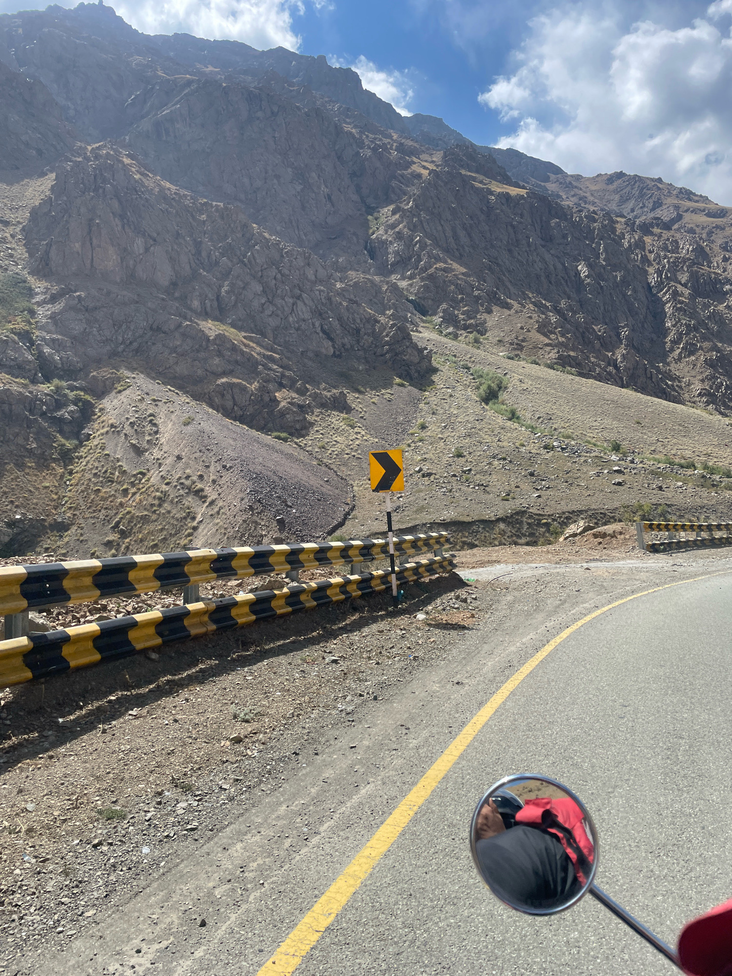 The image captures a scenic view of a mountainous region, likely a winding road in a remote area. The road itself is paved with a yellow line marking the lane. A guardrail with alternating black and yellow reflective stripes runs along the side of the road, separating it from the rough, rocky terrain. On the left side of the road, a signpost with a yellow chevron indicates a curve. The mountainous backdrop is characterized by steep, barren slopes with sparse vegetation. The sky above is partly cloudy, with bright white clouds against a blue sky. In the foreground, a motorcycle's mirror reflects the image of a person wearing a red jacket over a black shirt, suggesting the photo was taken from a moving vehicle.