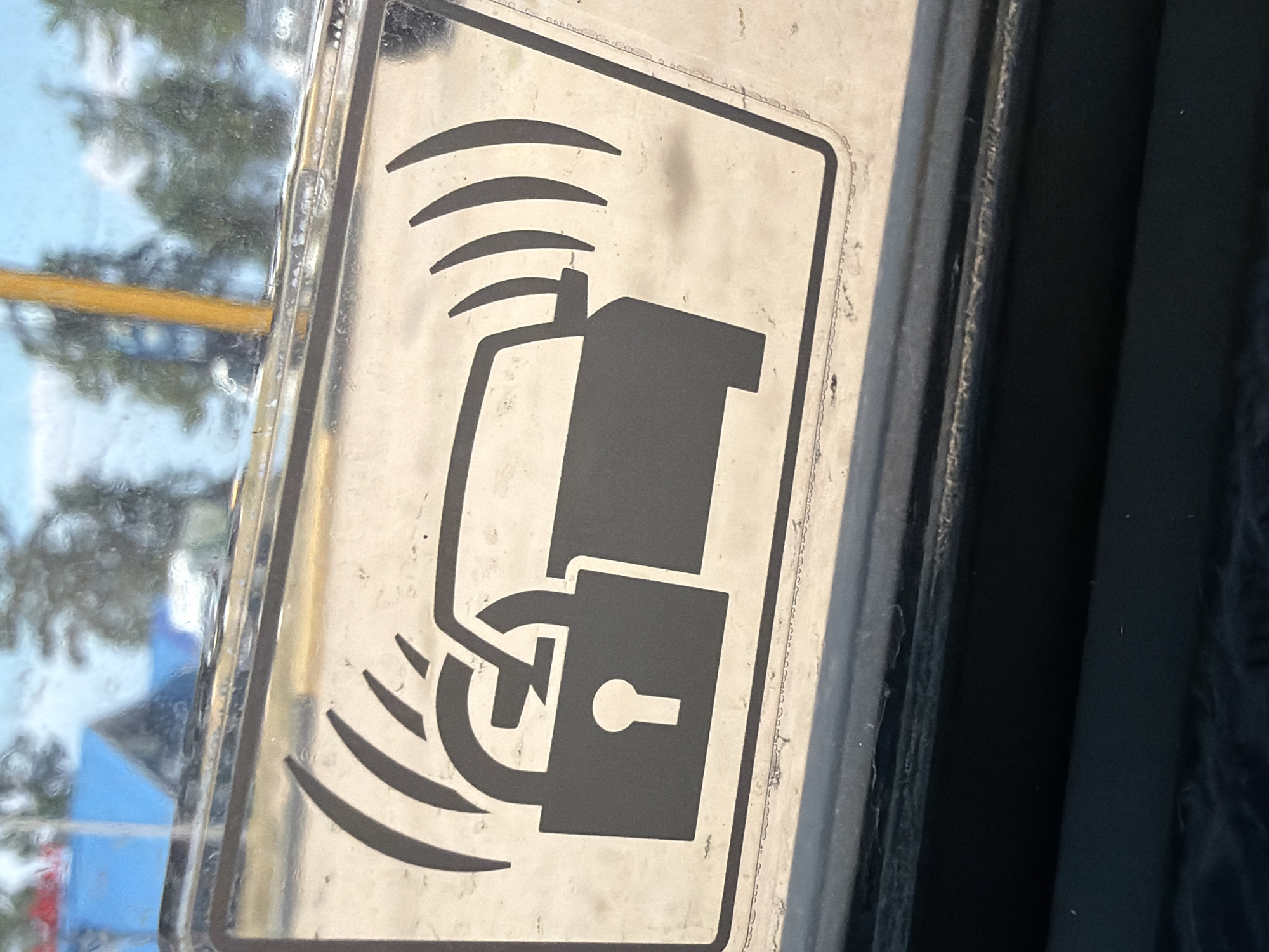 The image shows a sticker with a car alarm symbol on the interior of a vehicle's windshield. The symbol depicts a car with curved lines radiating from its rear, indicating a signal or alarm. To the left of the car is a padlock, also with radiating lines, suggesting it is also alarmed or secured. The sticker appears to be rectangular with a black border. The background, seen through the windshield, shows an outdoor environment with trees and a blue structure that could be a building. The image quality is somewhat blurred and affected by reflections on the glass. The sticker is located on the interior of the windshield of a vehicle. There are no people or specific actions depicted in the image beyond the static representation on the sticker. The environment outside appears to be a moderately sunny day with some foliage and structures visible. The specific location in the city of Turka, Russia, cannot be determined from the visual information provided in the image.