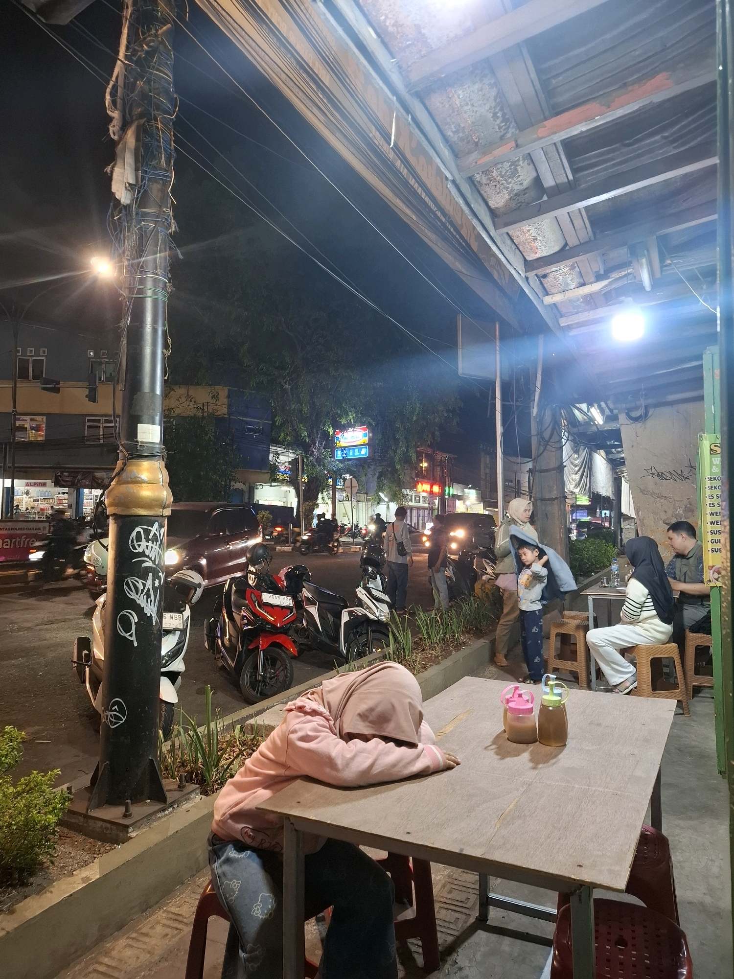 The image depicts a street-level view of a commercial area in Cianjur, Indonesia, at night. A central focus is a street light pole covered in wires and graffiti. To the right, a simple outdoor seating area of what appears to be a food stall or restaurant is set up under a rustic, open-air canopy. Several people are present: a person in a pink hoodie and hijab is slumped over a wooden table, seemingly asleep. Across the table, two individuals are seated, one wearing a hijab and the other a dark shirt. A child stands near them, partially covered by a blue garment.
In the background, the street is active with traffic, including parked motorcycles and cars. Buildings line the street, with visible signage indicating businesses. Trees with dense foliage are also present, contributing to the urban streetscape. The lighting suggests it is evening or night, with artificial lights illuminating the scene, creating areas of brightness and shadow. The overall impression is a bustling, everyday scene in an Indonesian city at night.