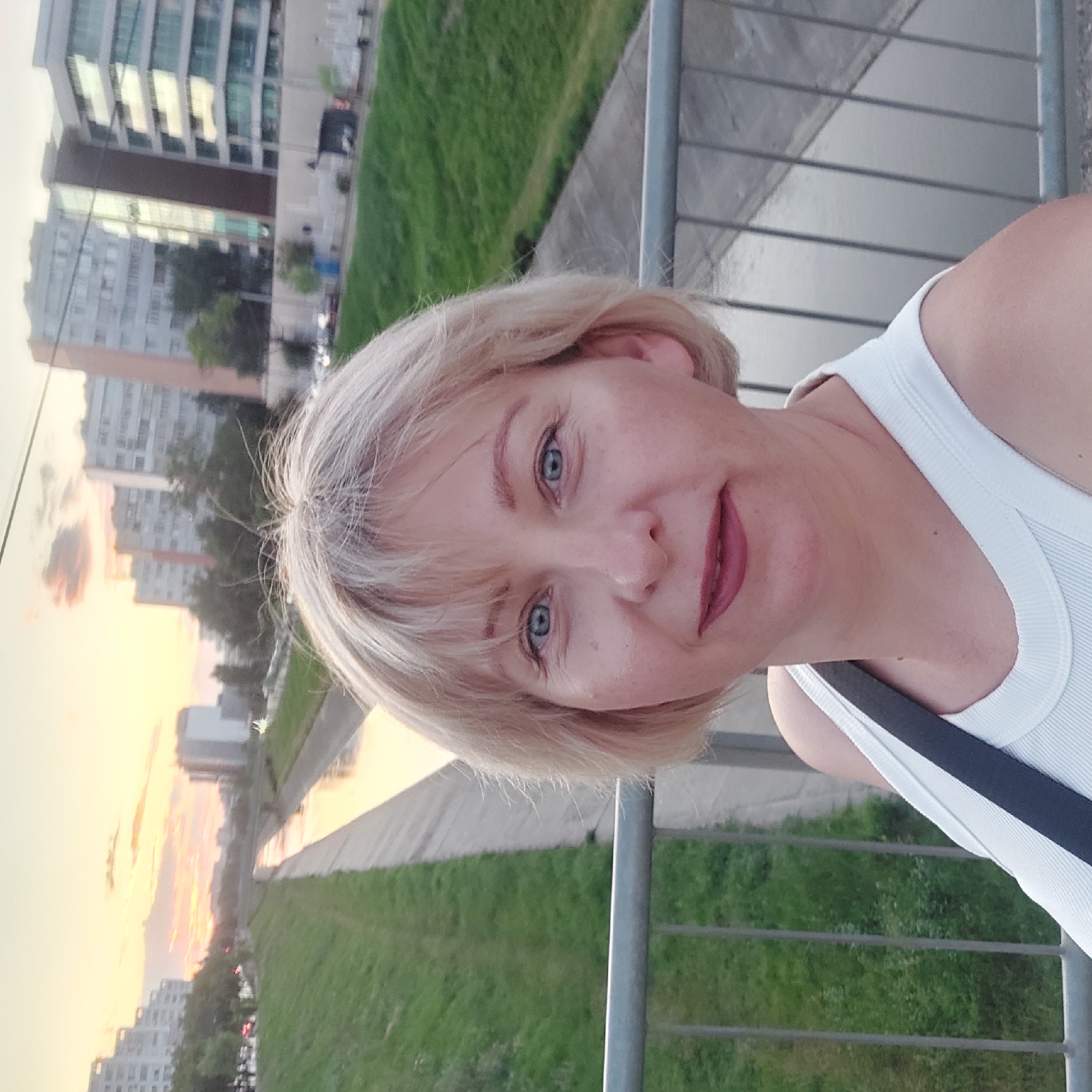 A selfie of a blonde woman with blue eyes and medium skin tone is taken on a bridge overlooking a canal in Iași, Romania, during sunset. The woman is wearing a white tank top. The canal, lined with concrete banks and grassy slopes, runs through a modern urban landscape with apartment buildings and trees visible in the background. The sky is painted with warm sunset colors.