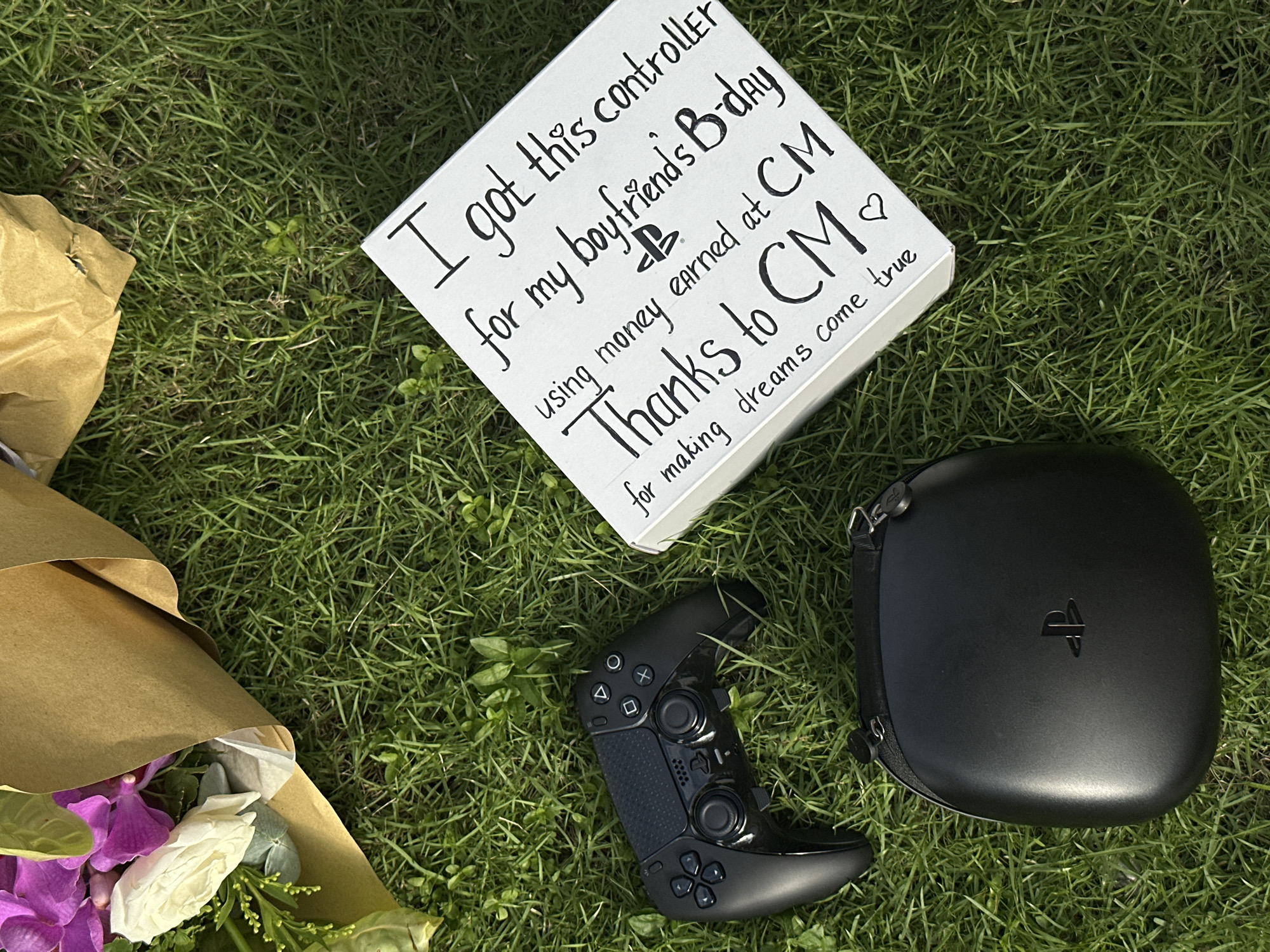 The image depicts a congratulatory display on a grassy surface. A sign with the text "I got this controller for my boyfriend's B-day using money earned at CM. Thanks to CM for making dreams come true" is centrally placed. Next to the sign is a black PlayStation controller and a case with a PlayStation logo. A bouquet of flowers wrapped in brown paper is visible on the left side of the frame. The exact location in North Kuta, Indonesia cannot be determined from the image.