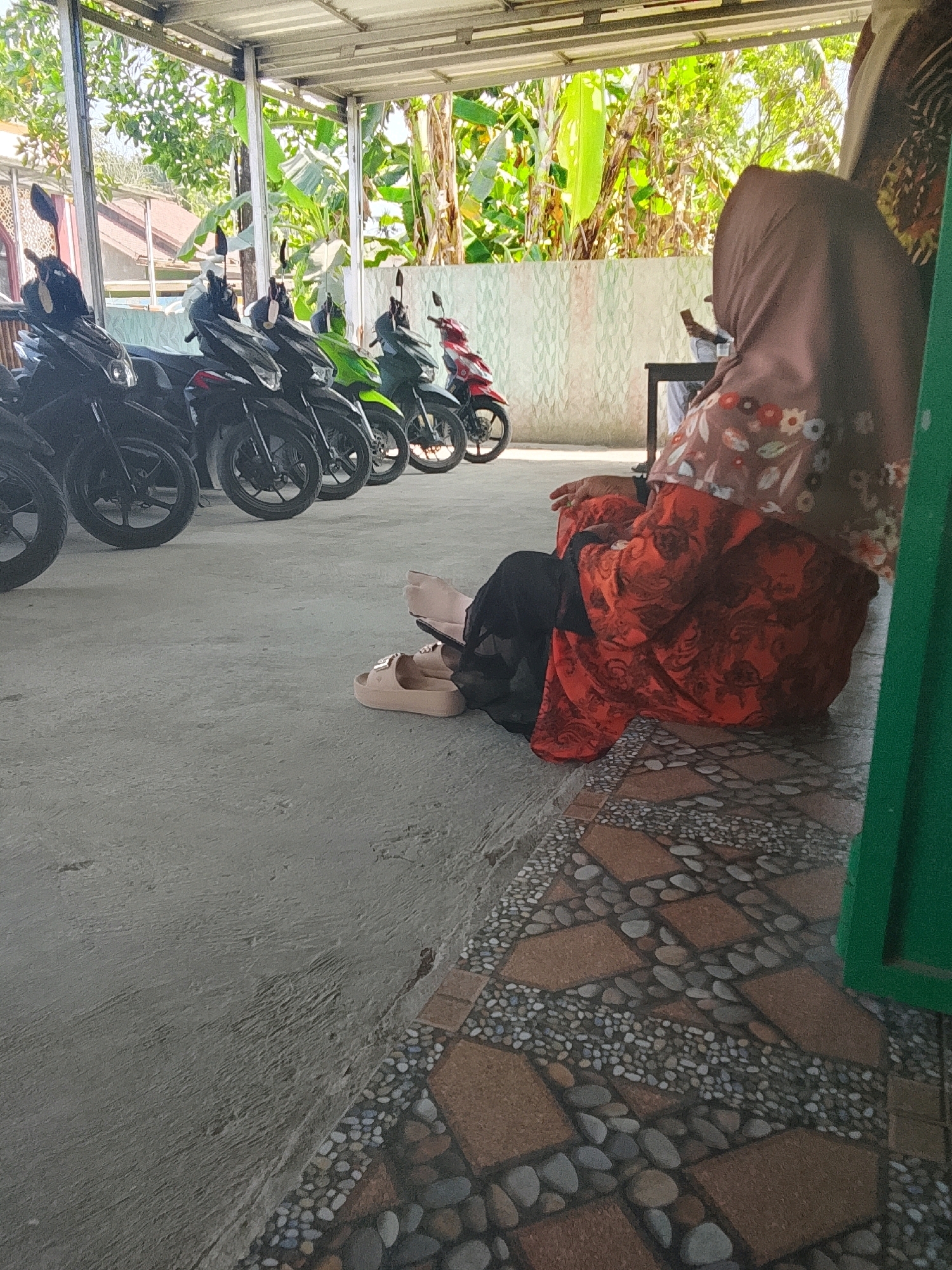 This image depicts an outdoor scene, likely in Tayem, Indonesia, under a covered structure that resembles a carport or an open-air market stall. Several motorcycles are parked in a row, suggesting a transportation hub or a place where people leave their vehicles. In the foreground, a person is seated, facing away from the camera, wearing a hijab and a long, patterned dress in shades of red and orange. Their legs are crossed, and a pair of light-colored sandals are placed on the ground near them. The ground is a mix of concrete and decorative tiling with a pebble mosaic pattern. In the background, lush green foliage and trees are visible, indicating a tropical or subtropical environment. The overall impression is one of a quiet, possibly everyday scene in a local Indonesian setting.