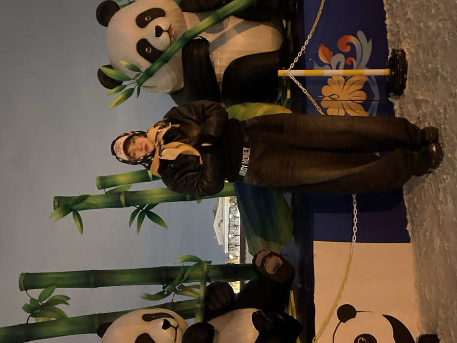 A person posing with panda-themed decorations. The decorations include a large panda figure with bamboo. The individual is wearing a dark jacket, jeans with the words "Daisy Honey" on the waistband, and a hat. A building with columns can be seen in the background. The setting appears to be outdoors, possibly in a public space with winter decorations. The image orientation is vertical.