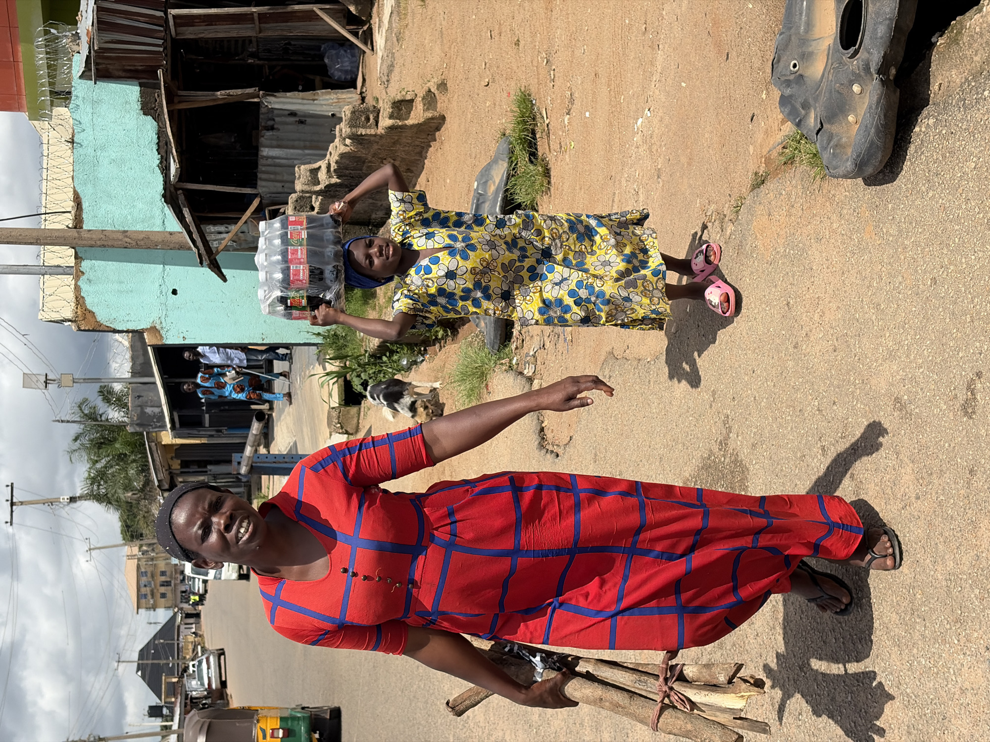 A full shot shows two women walking on a dusty road in Jos, Nigeria. The woman on the left is wearing a red dress with blue lines and carrying a bundle of sticks in her left arm. The woman on the right is wearing a yellow dress with blue flowers and carrying a tray of Coca-Cola bottles on her head. In the background, there are buildings and some vegetation. The sky is cloudy.