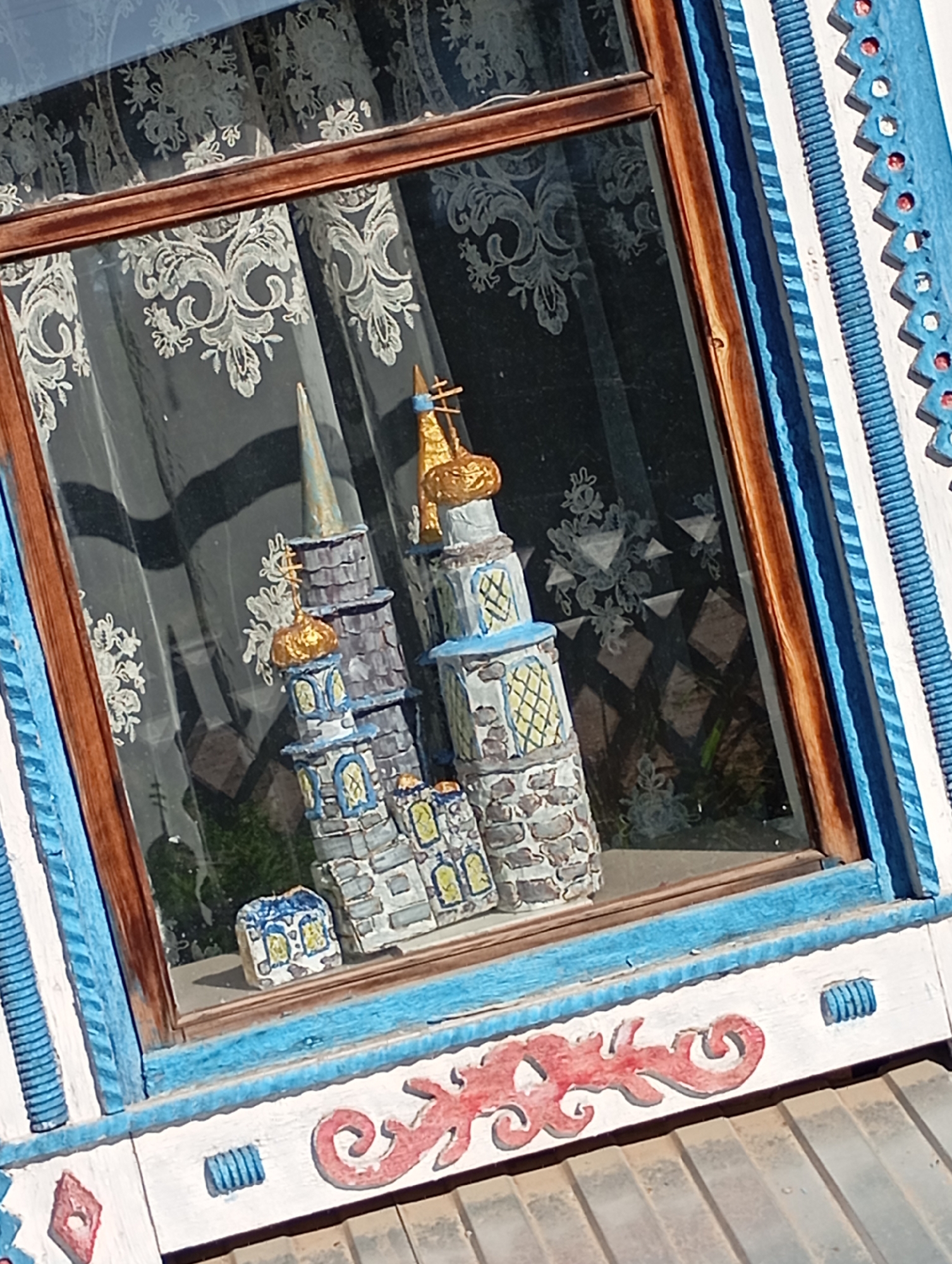 A low-angle shot captures a window, reflecting a display of miniature buildings against a backdrop of lace curtains and sunlight. The window frame features intricate blue and white carvings with red accents along the lower edge. The miniature buildings, resembling a castle, are painted in shades of gray, blue, and gold, with detailed windows and spires. The buildings sit on a windowsill. The location is in Unknown, Unknown.