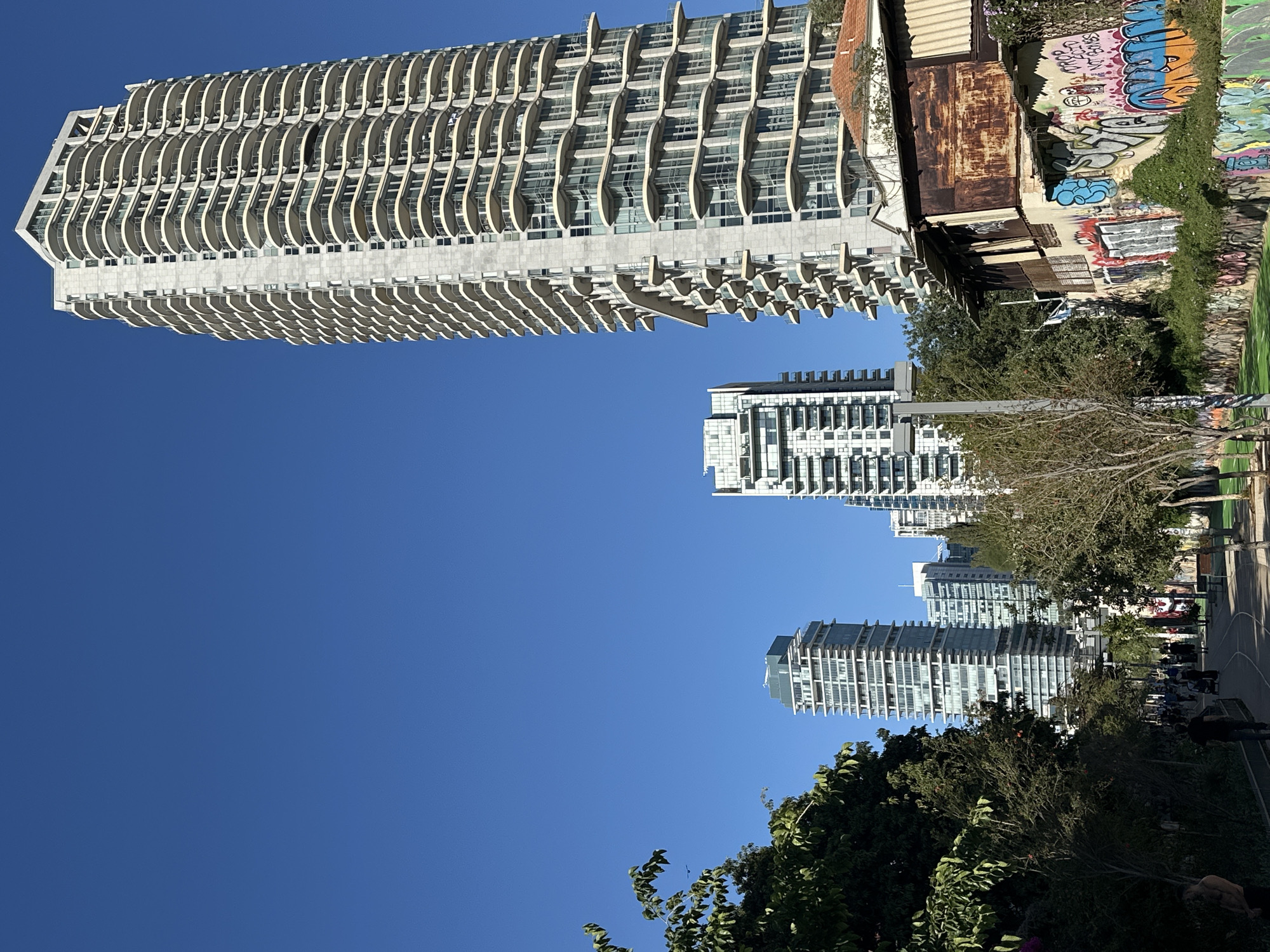 This image captures a vibrant urban scene in Tel Aviv, Israel, under a bright, clear blue sky.
Dominating the left side of the frame is a towering, modern high-rise building. It features a light-colored facade with numerous floors, each distinguished by a continuous, wave-like pattern of curved, elongated balconies or architectural elements.
Adjacent to this sleek structure, and creating a strong visual contrast, is a smaller, older building. This dilapidated structure features rusty, corrugated metal siding on its upper part and concrete or stucco walls below. These walls are extensively covered in colorful graffiti, with various tags and designs. Partial words like "ARMY" and "BLUE" are discernible within the intricate street art.
In the mid-ground and background towards the right, several other modern high-rise buildings stand, characterized by their glass and concrete construction. Below these buildings and extending across the right portion of the image is a public park or promenade. Lush green trees and dense foliage line a paved pathway where several people are visible, walking or standing, indicating leisurely activity.
The bright, direct sunlight suggests a clear and sunny day, likely mid-morning or afternoon. The overall scene presents a dynamic blend of contemporary architecture, urban grit, and public green spaces within a bustling city environment.