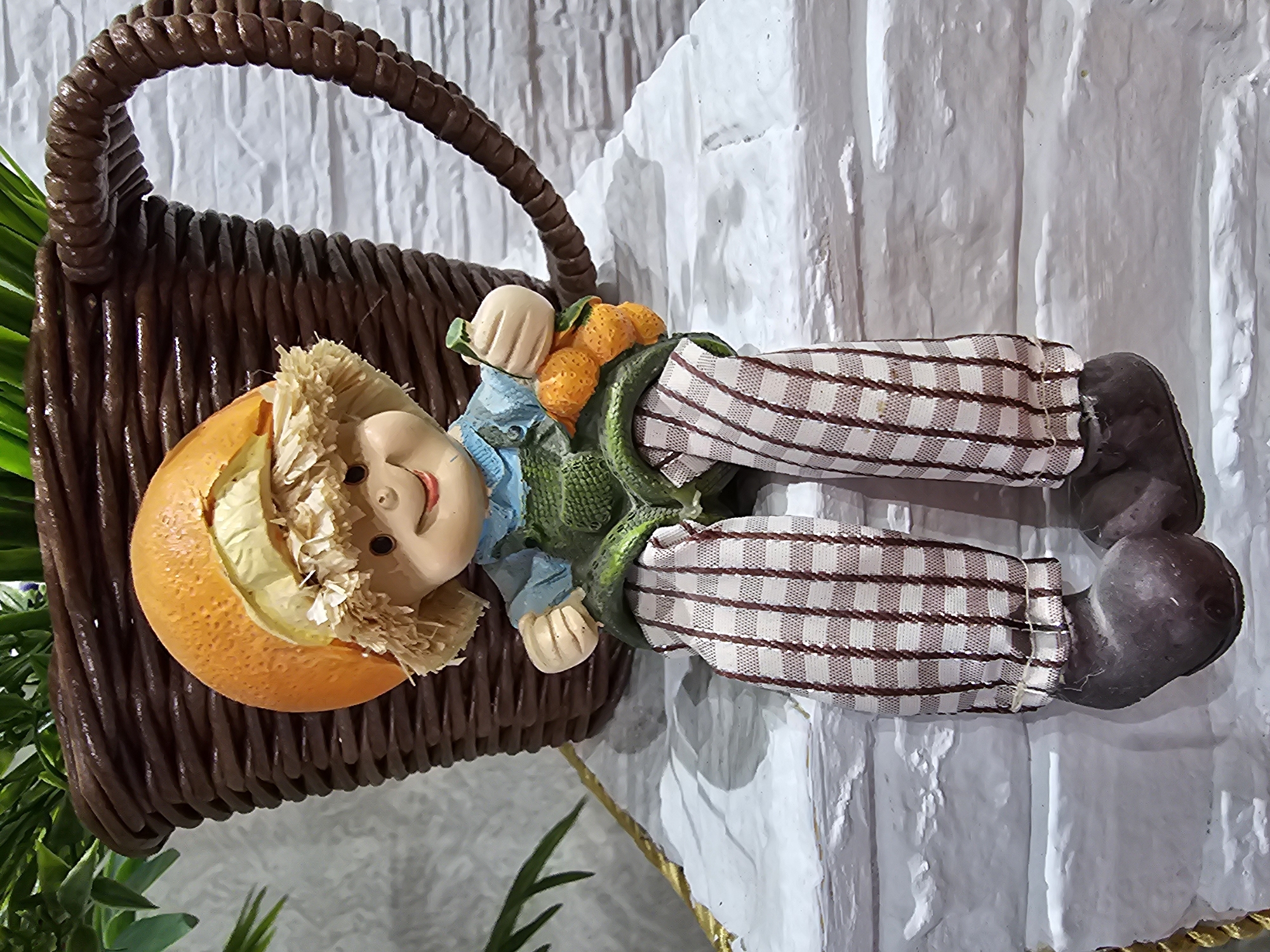 A garden gnome figurine is seated on a white brick ledge. The gnome has a straw hat made from a half orange, a blue shirt, green overalls, and brown and white checkered pants. It holds a cluster of small orange fruits and a green stem in its right hand. Behind the gnome is a large brown wicker basket, likely a planter, filled with green foliage. The background is a white brick wall. The image is taken at a medium distance, focusing on the figurine. The location is specified as Kalinovka, Russia.