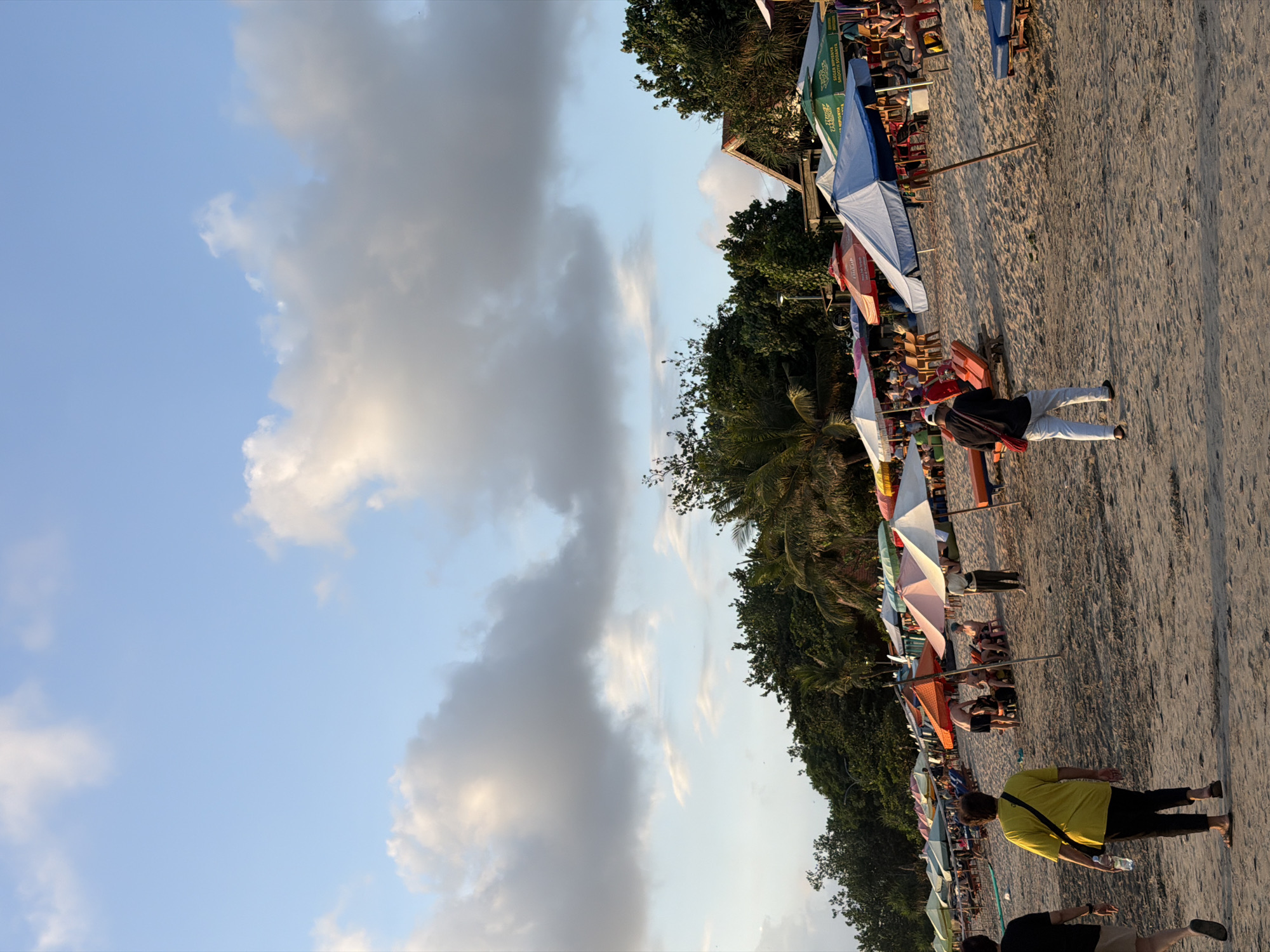 This image depicts a beach scene in Kuta, Indonesia, likely during the late afternoon or early evening given the warm lighting and long shadows. The sky is partly cloudy, with patches of blue sky interspersed with larger, gray clouds. The foreground is dominated by a sandy beach, where numerous beach umbrellas and chairs are set up. Some of these umbrellas are open, providing shade to what appear to be beachgoers, while others are closed.
Several people are visible on the beach. In the mid-ground, a person in a yellow shirt and black pants is walking towards the viewer, carrying a water bottle. Further back, another person in white pants and a dark top is walking away from the viewer. Several other individuals are seen sitting in chairs or walking near the umbrellas. The presence of many umbrellas and chairs suggests a popular tourist area.
Behind the beach umbrellas, there is a line of lush green trees and vegetation, with some buildings visible among the trees, hinting at the developed nature of the coastline. The overall atmosphere appears relaxed, consistent with a beach setting. No discernible text is visible in the image.