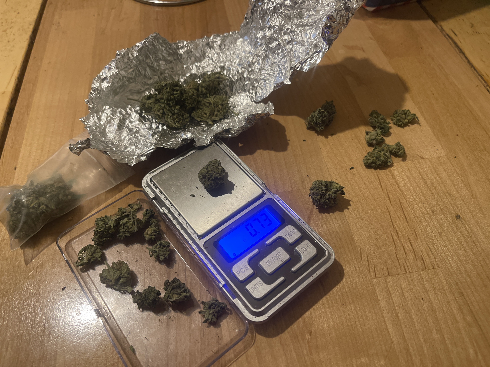 This image captures a close-up, high-angle view of green, leafy plant material, consistent with cannabis buds, being weighed and prepared on a wooden surface.
The central focus is a small silver digital scale with a bright blue backlit display showing "0.73 g". A single piece of the green plant material rests on the scale's platform. More of the plant material is scattered around the scene: a significant amount is contained within a crumpled piece of aluminum foil in the upper left, while several smaller buds are loose on the wooden surface to the right of the scale. A clear, small plastic bag, partially filled with the same material, lies to the left, and a clear plastic tray or lid holds additional pieces below the foil.
The wooden surface, likely a table or floor, has a light to medium brown tone with visible grain and minor wear. The lighting is artificial, casting soft shadows from the objects, suggesting an indoor setting.
Visible text on the digital scale includes "g" (for grams), "PCS", "UNITS", "ON/OFF", "TARE", and "LIGHT". The display clearly reads "0.73". The overall activity depicted is the measurement and organization of the plant material, possibly for personal use or preparation for distribution.