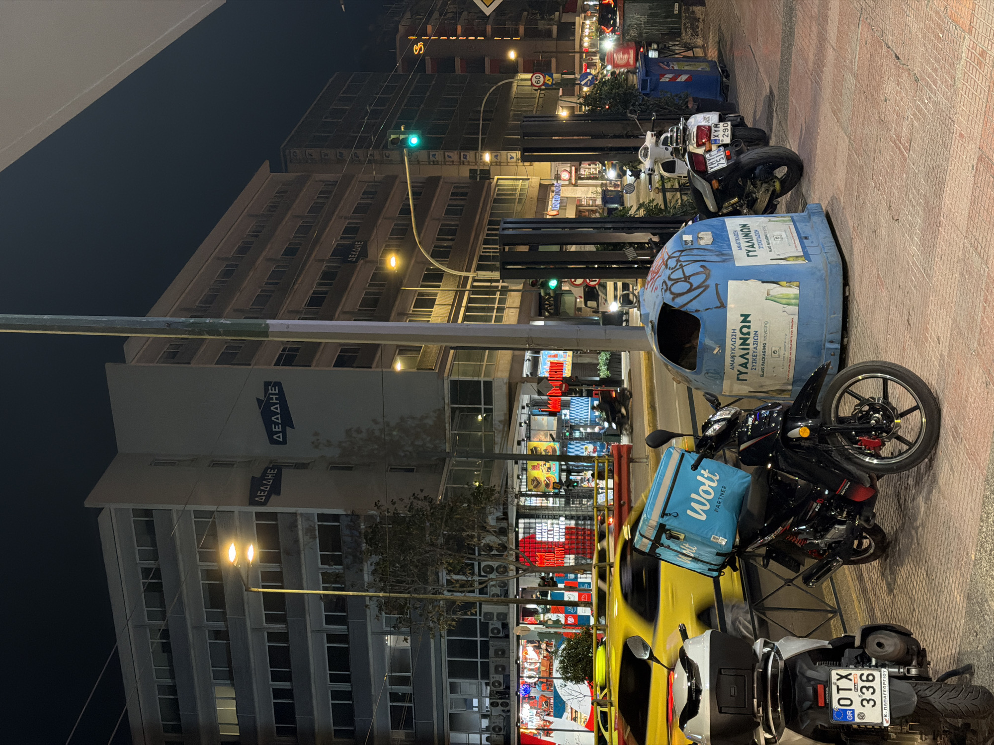The image captures a nighttime urban street scene, oriented as if viewed from a sidewalk, looking towards the street and buildings. Several motorcycles and scooters are parked on the paved sidewalk in the foreground, with a partial view of a yellow taxi beside one. A prominent blue recycling bin, clearly labeled "ΑΝΑΚΥΚΛΩΣΗ ΓΥΑΛΙΝΩΝ ΣΥΣΚΕΥΑΣΙΩΝ" (Glass Packaging Recycling) and "GLASS PACKAGING recycling," stands near the parked vehicles. One of the scooters is equipped with a blue "Wolt PARTNER" delivery bag, suggesting commercial activity.
Across the street, multi-story buildings dominate the background, with several windows illuminated. The Greek acronym "ΔΕΔΔΗΕ" (DEDDIE), belonging to the Hellenic Electricity Distribution Network Operator, is visible on at least two of these buildings. Ground-floor shops display various illuminated signs and products, casting bright colors onto the scene. A green traffic light is visible mid-frame, indicating an active road. Streetlights provide general illumination, and the sky is dark, confirming the nighttime setting.
Location cues include the Greek language on signs and the "GR" country code on visible license plates (e.g., "GR OTX 336" with "Κ. ΠΑΠΑΓΕΩΡΓΙΟΥ," and "XYM 1516 290"). A speed limit sign of "60" is also present. The overall atmosphere is that of a bustling city evening in Athens, Greece, with typical urban infrastructure and delivery services in operation. The weather appears clear.