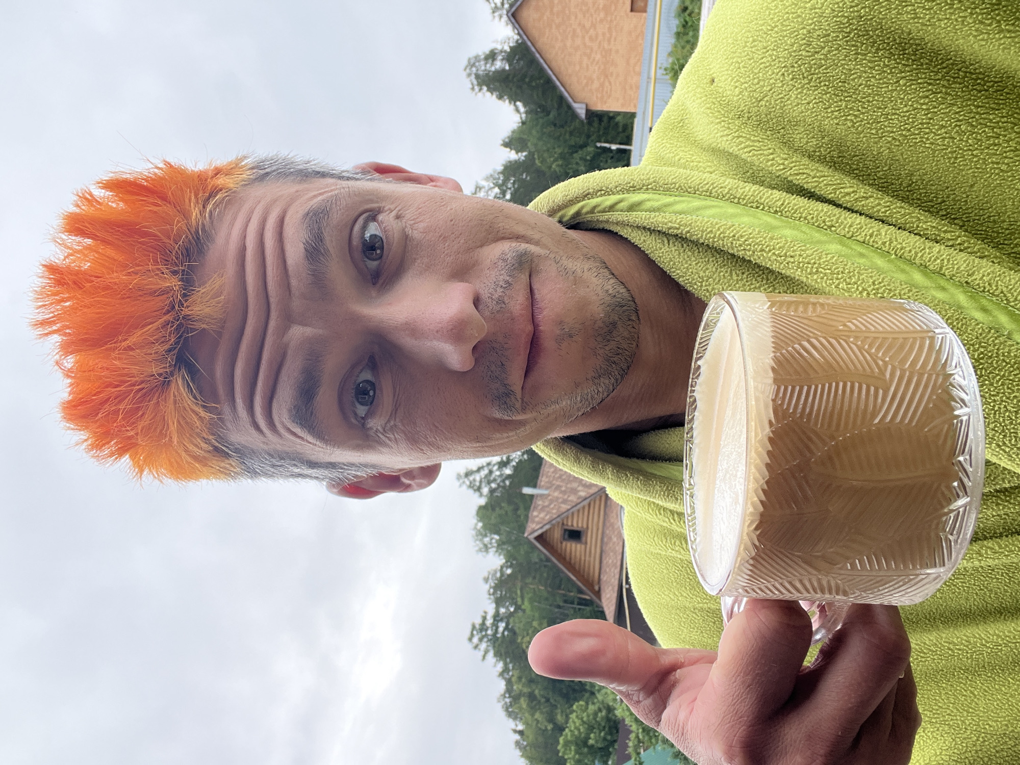 In Chaikovsky, Russia, a man with bright orange, spiky hair and a green robe is holding a clear glass mug filled with coffee. He is giving a thumbs-up to the camera. The background shows a rural landscape with trees and houses under an overcast sky.