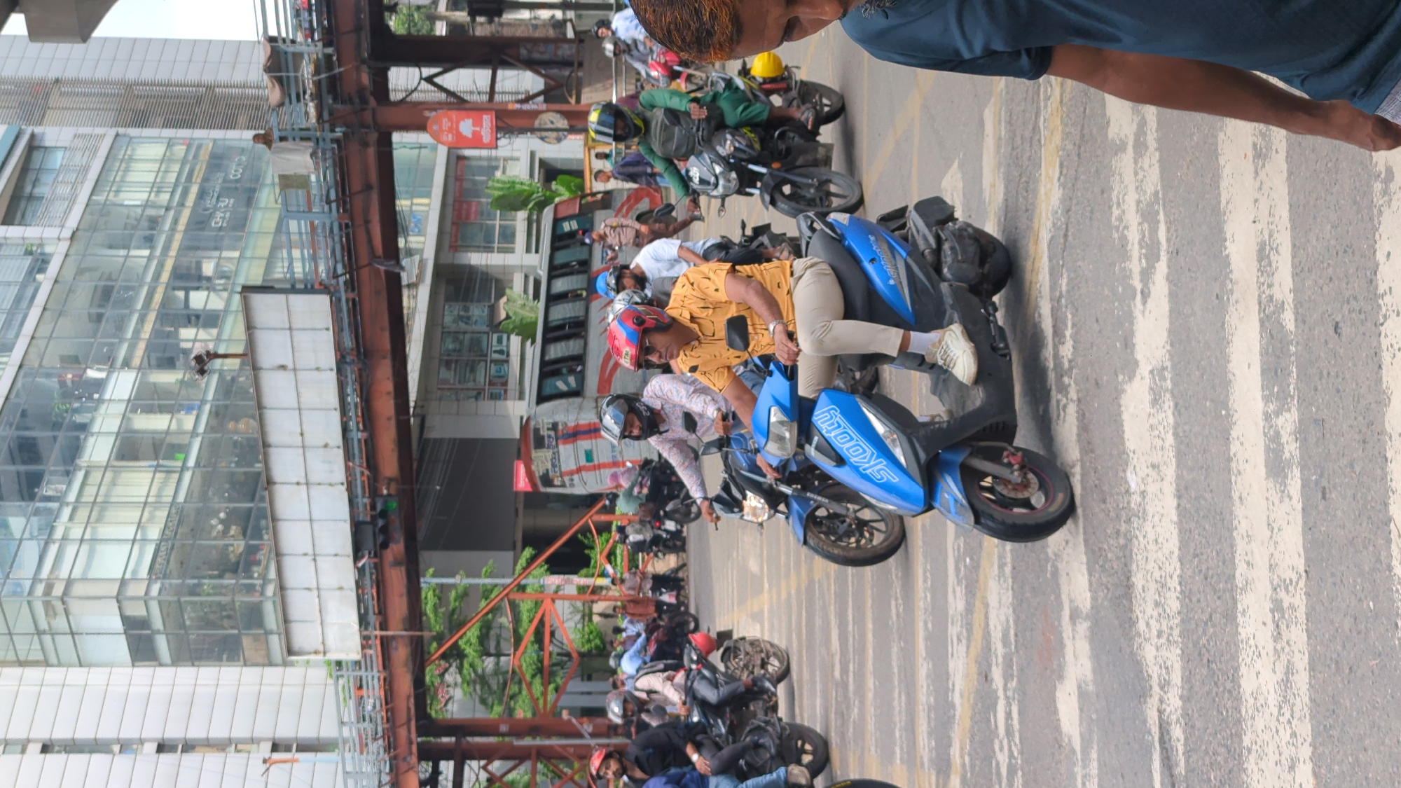 The image captures a street scene in Dhaka, Bangladesh, characterized by a busy intersection with several individuals riding motorcycles and scooters. In the foreground, a man in a yellow shirt and beige pants is riding a blue scooter. He is wearing a red and blue helmet and sunglasses. Behind him, other riders on motorcycles, some wearing helmets and some without, are visible. The background features a large, modern glass-fronted building and an elevated walkway or bridge structure. The road surface shows a pedestrian crossing. The overall atmosphere suggests an urban environment with active transportation.