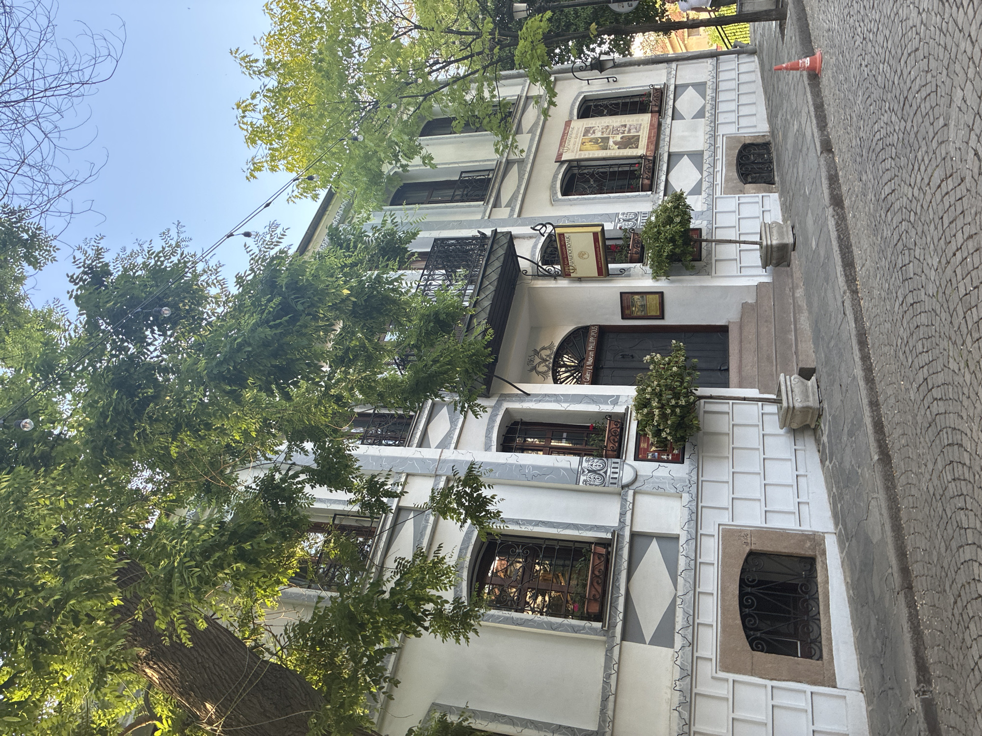 A low-angle shot reveals a white building with ornate detailing, including geometric patterns and decorative window frames. The architecture suggests an older style. A large tree with green foliage partially obscures the building's facade. Streetlamps and decorative elements are visible among the branches. A cobblestone street is on the right. The image is likely taken in the city of Plovdiv, Bulgaria.
