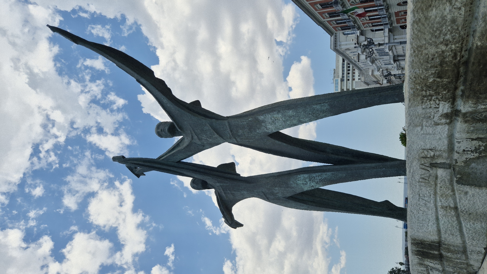 A low-angle, wide shot captures the "Monumento ai Caduti" (Monument to the Fallen) in Taranto, Italy, against a backdrop of a blue sky with scattered white clouds. The monument, made of dark bronze, depicts two stylized, abstract human figures, presumably male, standing close together. They are holding a large, draped fabric or banner aloft with their outstretched arms, suggesting a gesture of victory, defiance, or commemoration. The figures are elongated and angular, with simple, defined facial features. The monument rests on a large, rough-hewn stone base. Inscriptions are visible on the front of the stone base, partially obscured and worn by time.
In the background, a row of classical European-style buildings with ornate facades lines a street. The buildings are of varying heights and architectural styles, with balconies, arched windows, and street lamps. A flag, possibly the Italian flag with its green, white, and red stripes, hangs from one of the buildings. Further in the distance, a modern, multi-story building stands out against the sky. A body of water, likely the sea, is visible beyond the monument, with a few trees and distant landmasses on the horizon. The overall scene conveys a sense of historical significance and civic pride.