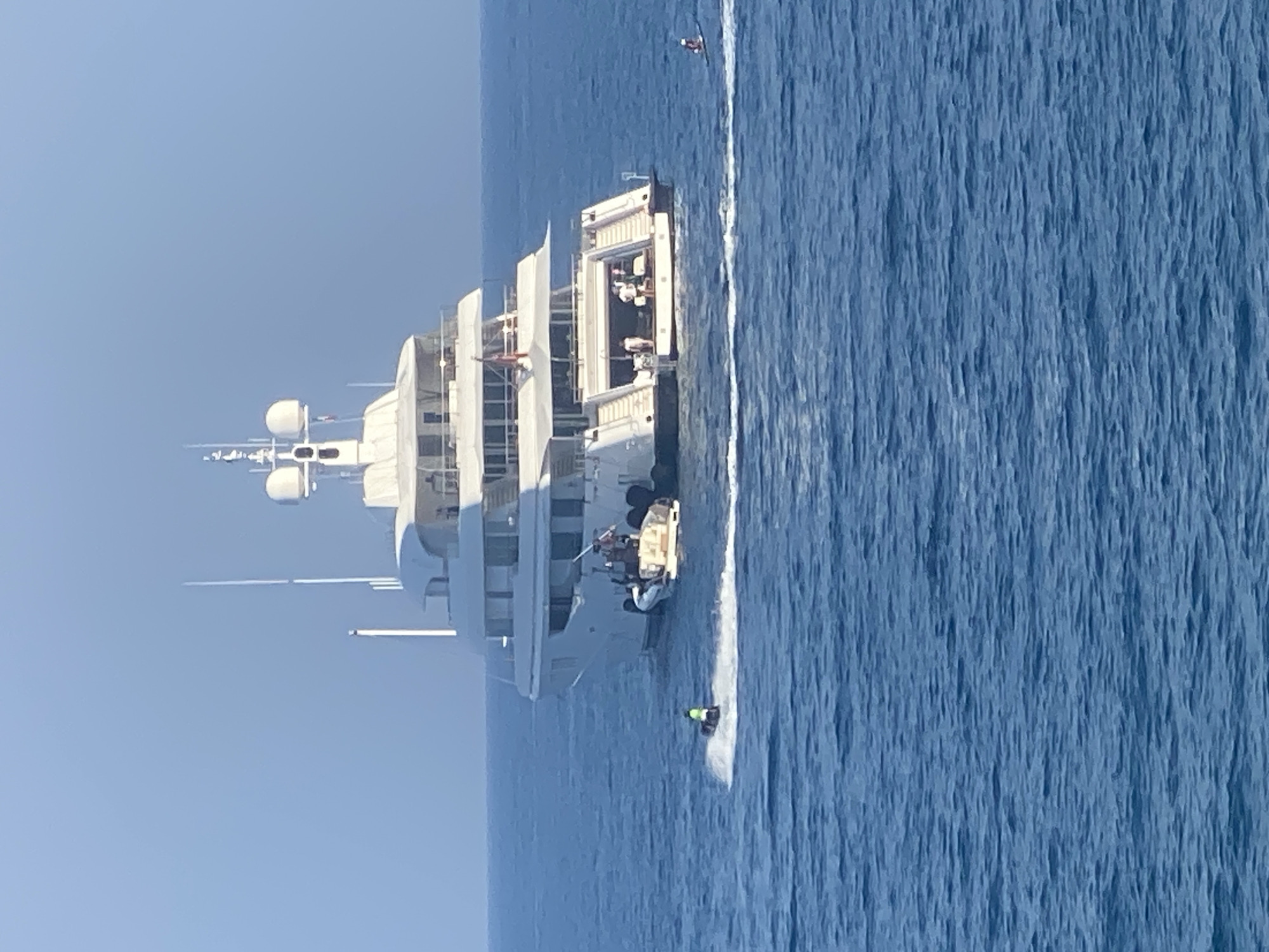 A large, white luxury yacht is anchored in the blue ocean off the coast of Antibes, France. The yacht has multiple decks and a tender boat is attached to the stern. On the main deck, several people are visible standing near the stern. A person is riding a jet ski in the foreground to the left, leaving a white wake behind them. In the distance to the right, another person is visible on a paddleboard. The sky is clear and blue.