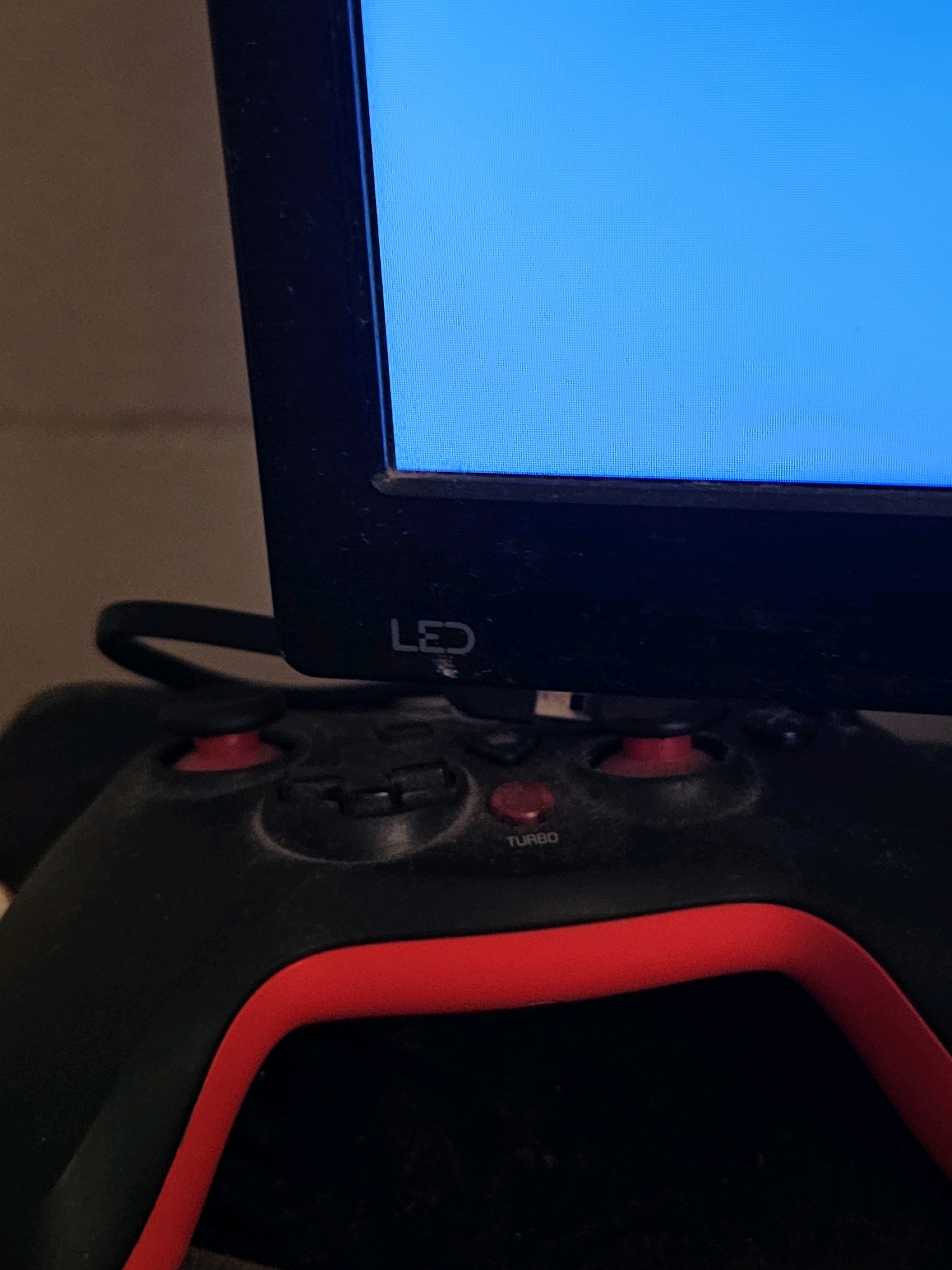 The image displays a close-up of a black video game controller with red accents, positioned beneath a monitor screen showing a bright blue display. The controller has several buttons visible, including one labeled "TURBO." The monitor has a black bezel, and text that appears to read "LED" is visible on the bezel. The background is a plain, light brown wall, suggesting an indoor domestic setting, potentially a living room or gaming area. There is visible dust on the controller and the monitor's bezel. There are no people present in the image. The scene is static and does not depict any action. Given the lack of distinguishing urban features, it is impossible to pinpoint a specific location within the city of Batna, Algeria.