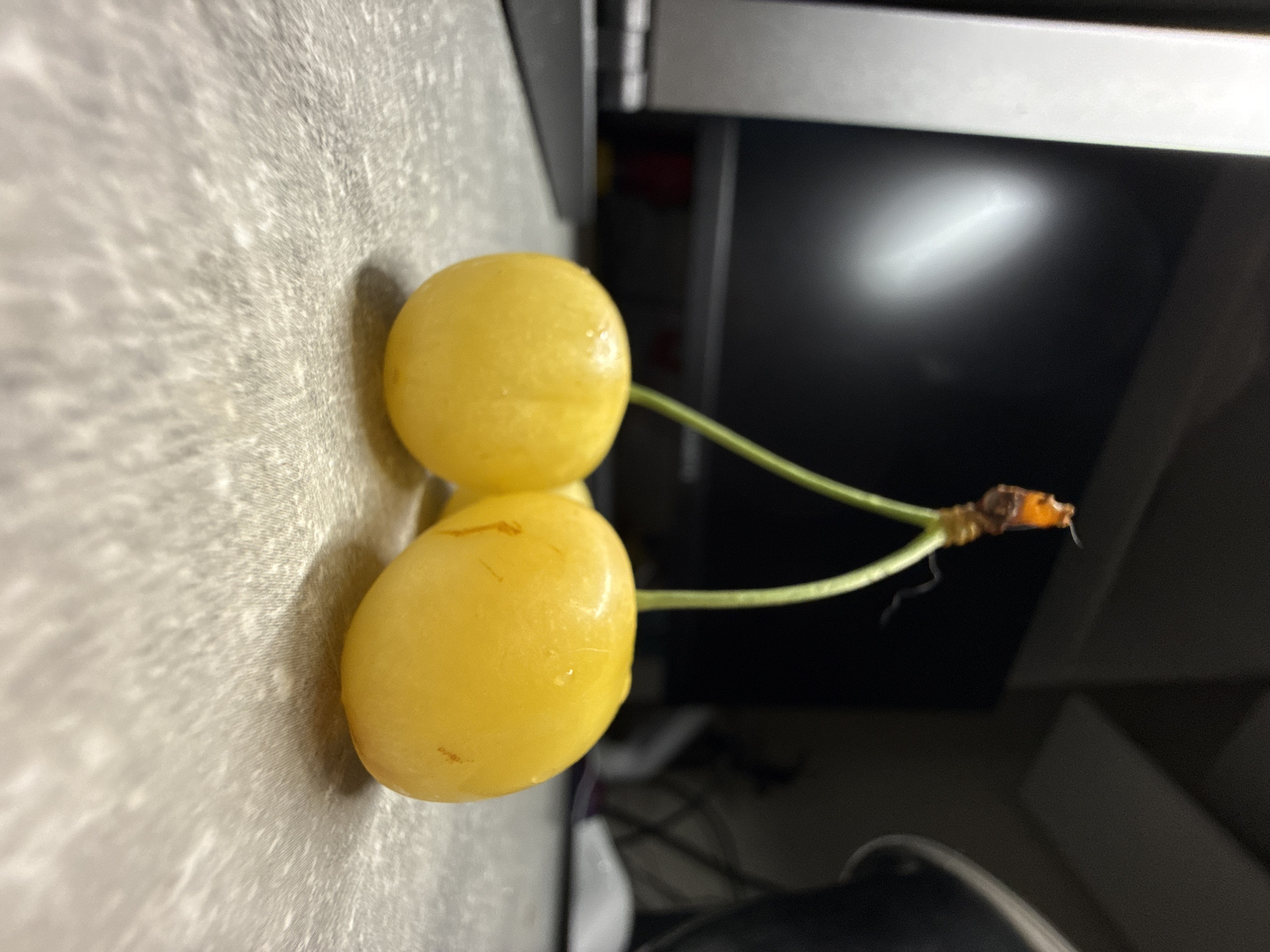 Two yellow cherries with stems are resting on a gray, textured surface. The cherries are positioned close to each other, with their stems reaching upwards and joining at a single point. The background is blurred, but appears to show a dark computer monitor and possibly some kitchen elements. The lighting is dim, creating shadows and highlighting the sheen of the cherries.