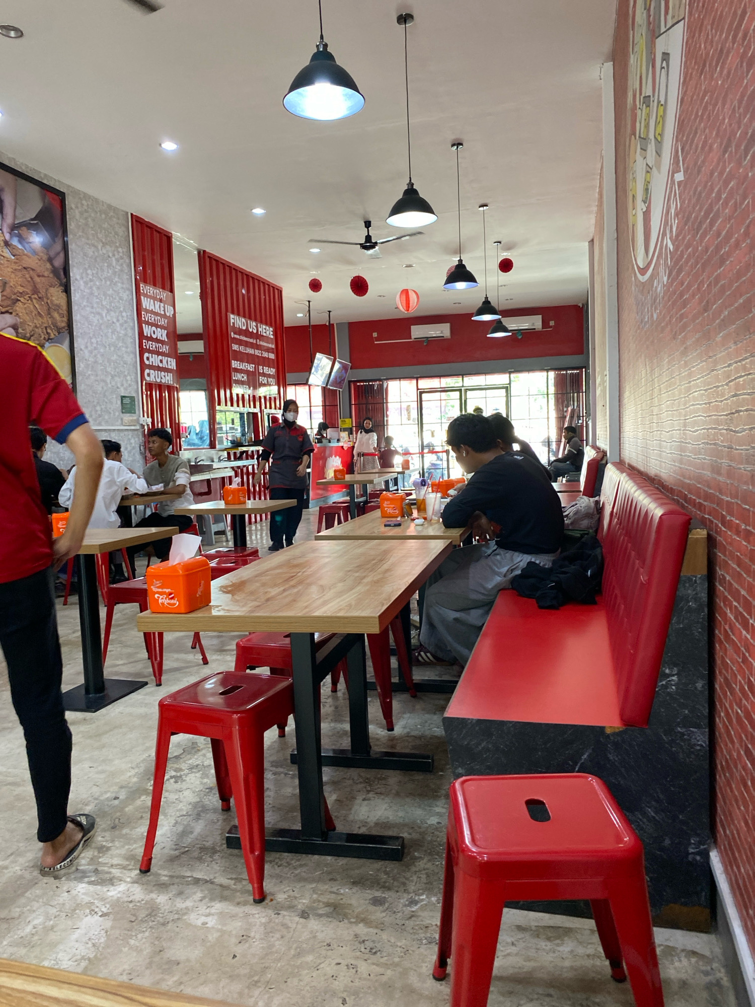 The image shows the interior of a restaurant in Banjarbaru, Indonesia, characterized by its red and white color scheme and minimalist design. The lighting fixtures are industrial-style pendant lamps. Red tables and chairs are arranged throughout the dining area, with some patrons seated and others standing. A red booth seating area lines the right wall. The walls are adorned with red brick patterns and motivational posters, one of which reads "EVERYDAY WAKE UP EVERYDAY WORK EVERYDAY CHICKEN CRUSH!". A counter area is visible in the background, where a staff member in a uniform is interacting with customers. The overall atmosphere suggests a casual dining establishment.
