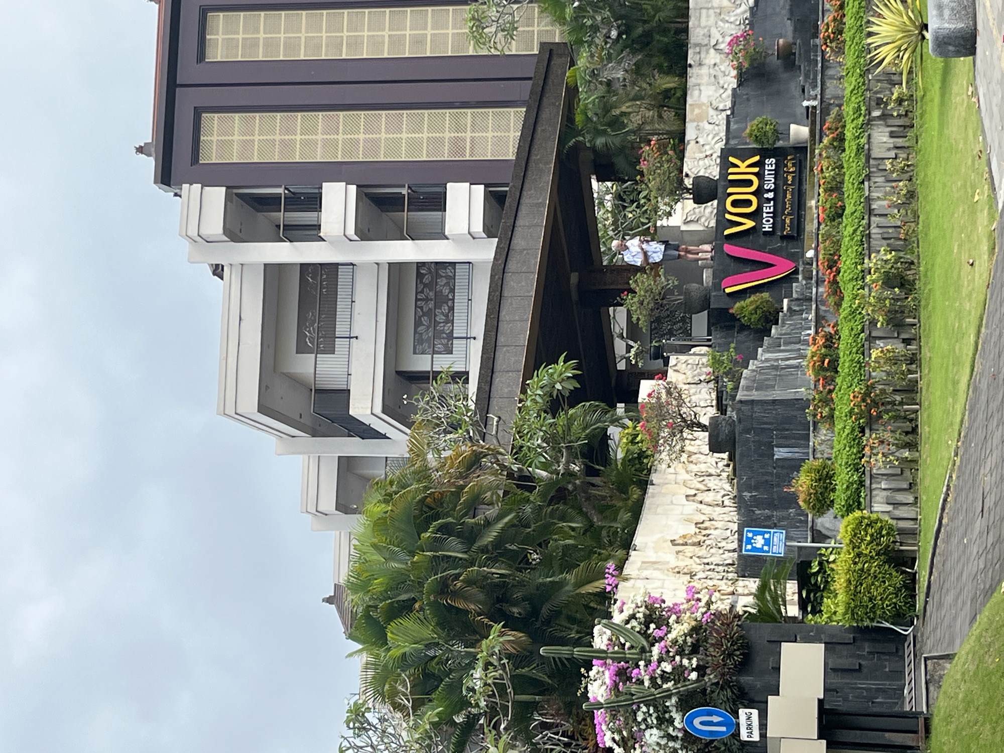 In Kuta Selatan, Indonesia, the VOUK Hotel & Suites is depicted with its prominent signage. The hotel features a modern architectural design with multiple levels and balconies. In front of the hotel, there is a landscaped area with various plants, including palm trees and flowering bushes. A man is standing on a raised platform near the entrance, looking at his phone. The scene is set on an overcast day, with a pathway leading towards the hotel entrance. Signs indicate parking and pedestrian access.