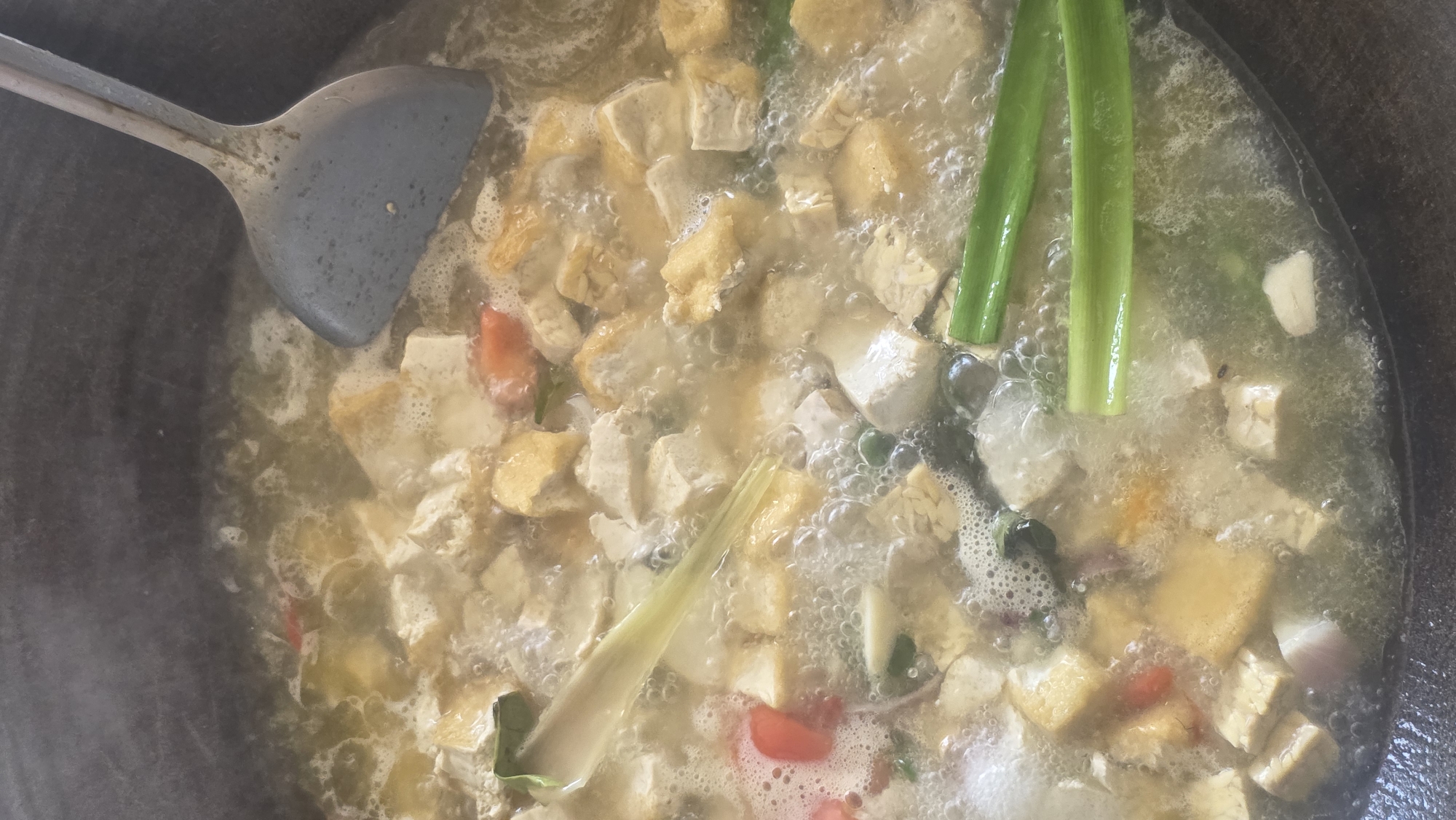 The image shows a top-down view of a simmering dish, likely a soup or stew, inside a dark-colored pot. A metal spatula rests on the edge, partially submerged in the liquid. The dish contains cubed tofu and possibly tempeh, as well as green stalks (likely scallions or lemongrass), and red pieces, possibly tomatoes or chilies. The broth is bubbling and appears slightly opaque. The context of the photo, if it was taken in Lhokseumawe, Indonesia, suggests this could be a local dish being prepared.