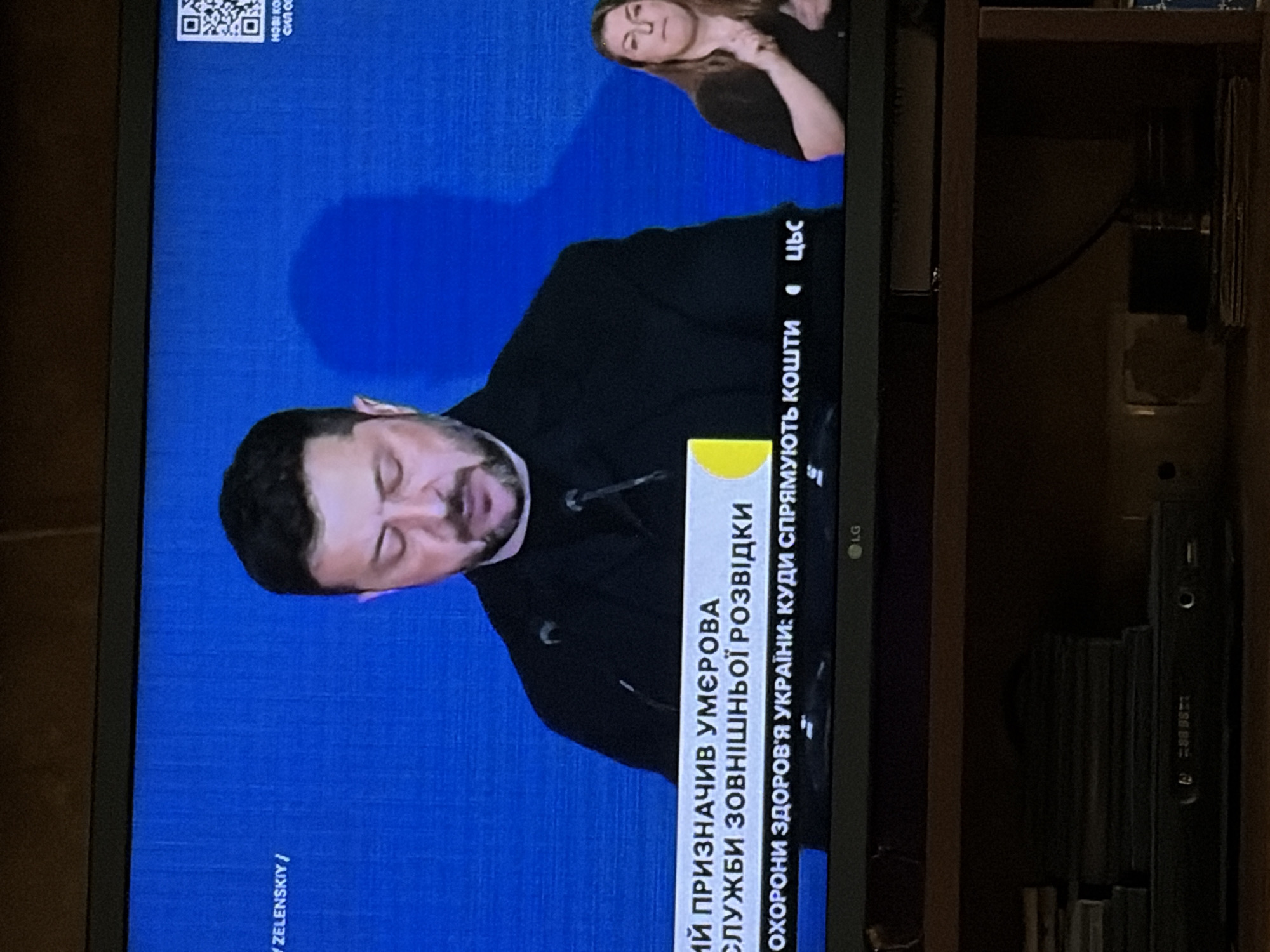 The image shows a television screen displaying a man, identified as Zelenskyy, speaking at a podium. He is wearing a black shirt and has a beard. There is a woman on the right side of the screen, also visible and appearing to be using sign language. The background is a solid blue screen with text overlays. One text overlay in the lower portion of the screen reads "ОХОРОНИ ЗДОРОВ'Я УКРАЇНИ: КУДИ СПРЯМ". The LG logo is visible at the bottom of the television screen. The television is placed on a shelf above other electronic devices and stacked media.