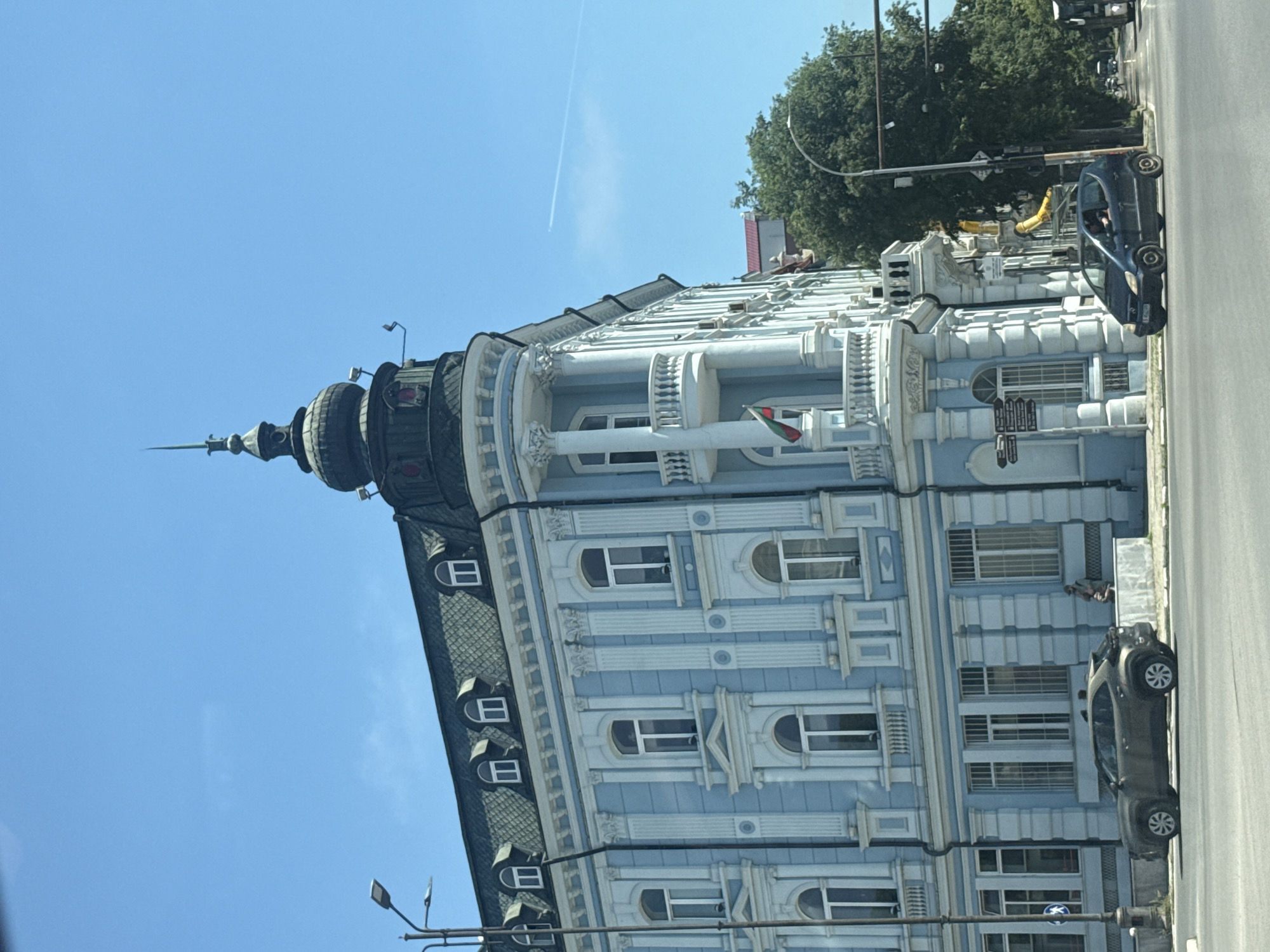 The image shows a view of the central part of Varna, Bulgaria, specifically on a street corner. The focal point is a large, ornate light blue building with white trim and architectural details, including columns and balconies. The building has a distinctive domed tower with a spire on the corner. A Bulgarian flag is visible on a balcony.
In the foreground, there are two cars driving on a paved road. The car on the left is a dark gray SUV, and the car on the right is a dark blue hatchback. Several street lights and traffic signals are present. Behind the building, lush green trees are visible. The sky is clear and blue with a faint contrail from an airplane. A single person is sitting on a ledge near the building, and another person is standing in front of the building. The overall impression is a sunny day in a bustling urban environment.