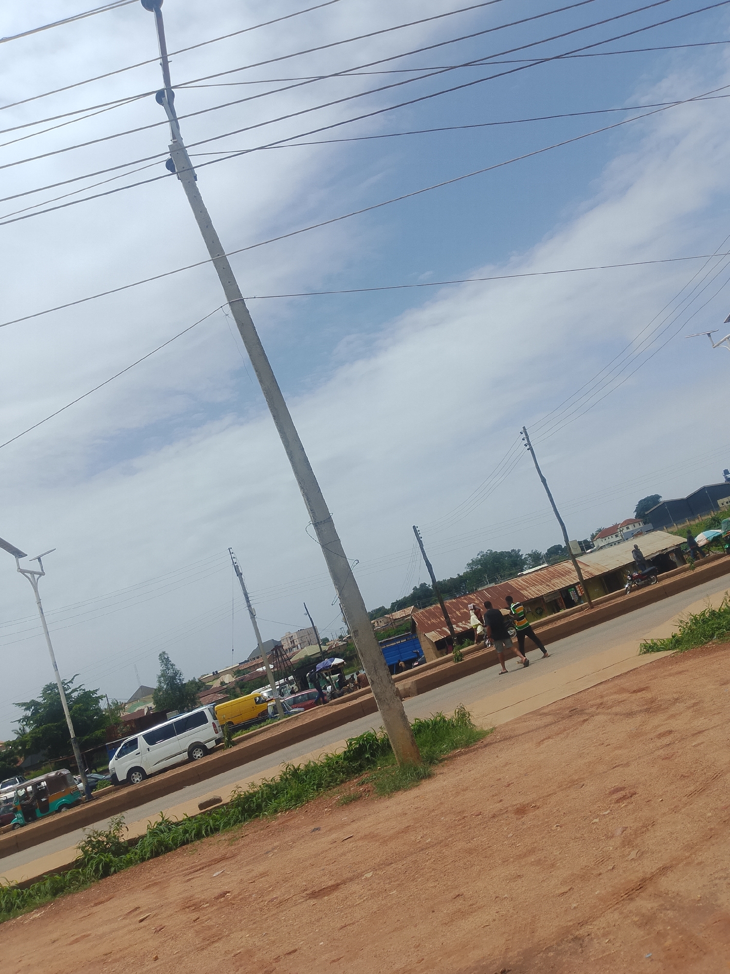 The image captures a street scene in Bukuru, Nigeria, under a partly cloudy sky, suggesting it is daytime. A prominent utility pole with multiple wires runs vertically through the frame, dominating the foreground. The scene depicts a road with several vehicles, including a white minivan, a yellow bus, a blue truck, and a tricycle.
In the background, there are buildings with corrugated iron roofs, indicative of a commercial or residential area. Two individuals are walking on the side of the road, one wearing a green and yellow striped shirt and the other in dark clothing. The ground is a mixture of dirt and sparse vegetation, with a paved road running through the middle of the frame.
The overall impression is of a typical day in a busy, possibly developing, urban or semi-urban environment. The lighting suggests it is not early morning or late evening. No visible text is present in the image.