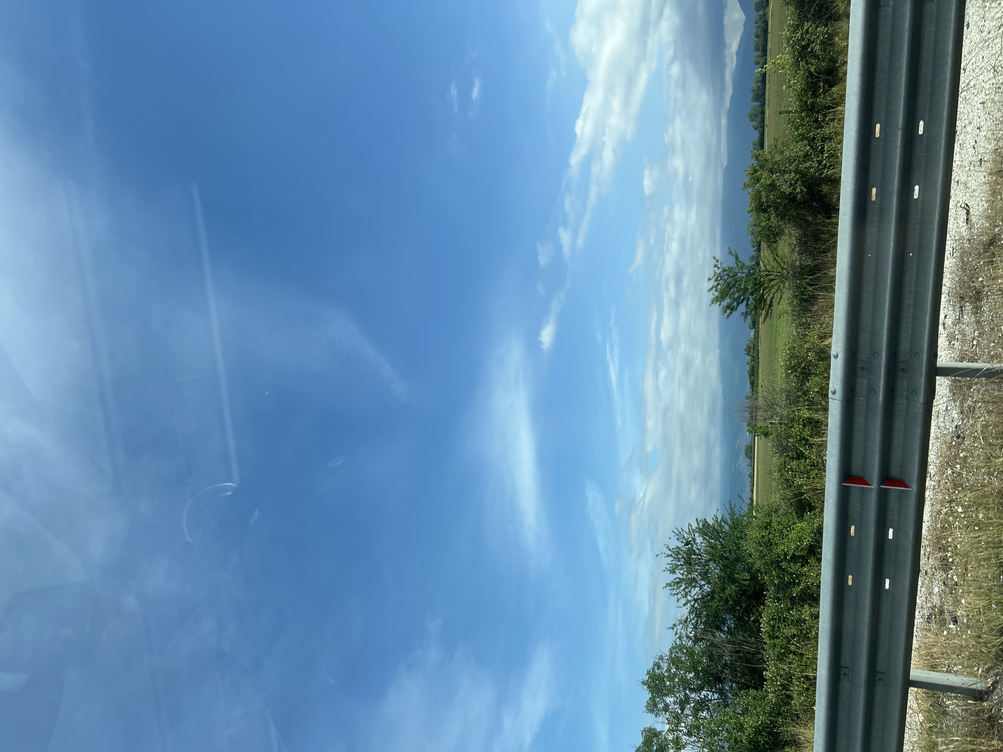 The image captures a bright, sunny daytime scene, likely viewed from a moving vehicle in Malo Konare, Bulgaria. The upper left portion of the frame shows a clear blue sky with some wispy white clouds and light reflections, possibly from a car window. Moving across the sky to the right, more prominent cumulus clouds are scattered, particularly towards the horizon.
Below the sky, the landscape features extensive green fields, indicative of agricultural land, dotted with various trees and bushes. In the distance, a range of mountains or hills with a hazy blue hue forms the backdrop, partially obscured by clouds.
In the immediate foreground, occupying the right side of the image, is a silver metal guardrail, commonly found alongside roads or highways. It runs horizontally across the frame, showing its corrugated structure and several mounting bolts. Two red reflectors are visible on the guardrail, and additional white reflective markings are present on its surface. The ground adjacent to the guardrail appears to be gravelly or dirt. The overall impression is one of a scenic journey through a rural landscape. No visible text is present.