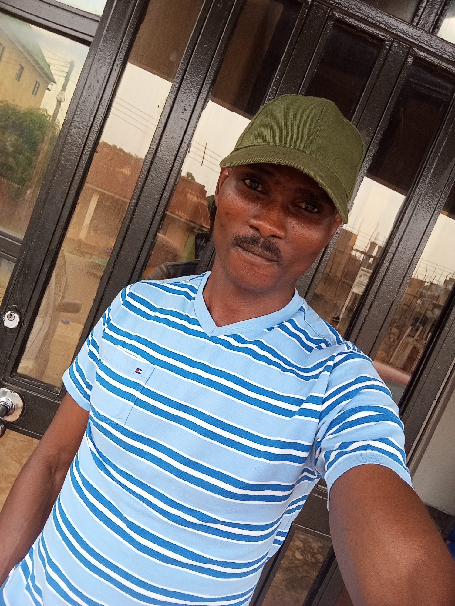 A close-up, eye-level, indoor selfie shows a dark-skinned man wearing a green baseball cap and a blue and white horizontally striped polo shirt. He has a mustache. He is standing in front of a dark-colored, glass-paned door with a metal frame. Reflections in the glass show buildings and trees, suggesting an urban or suburban setting. A round, silver doorknob is visible on the left side of the door. The image is well-lit, possibly by natural light.