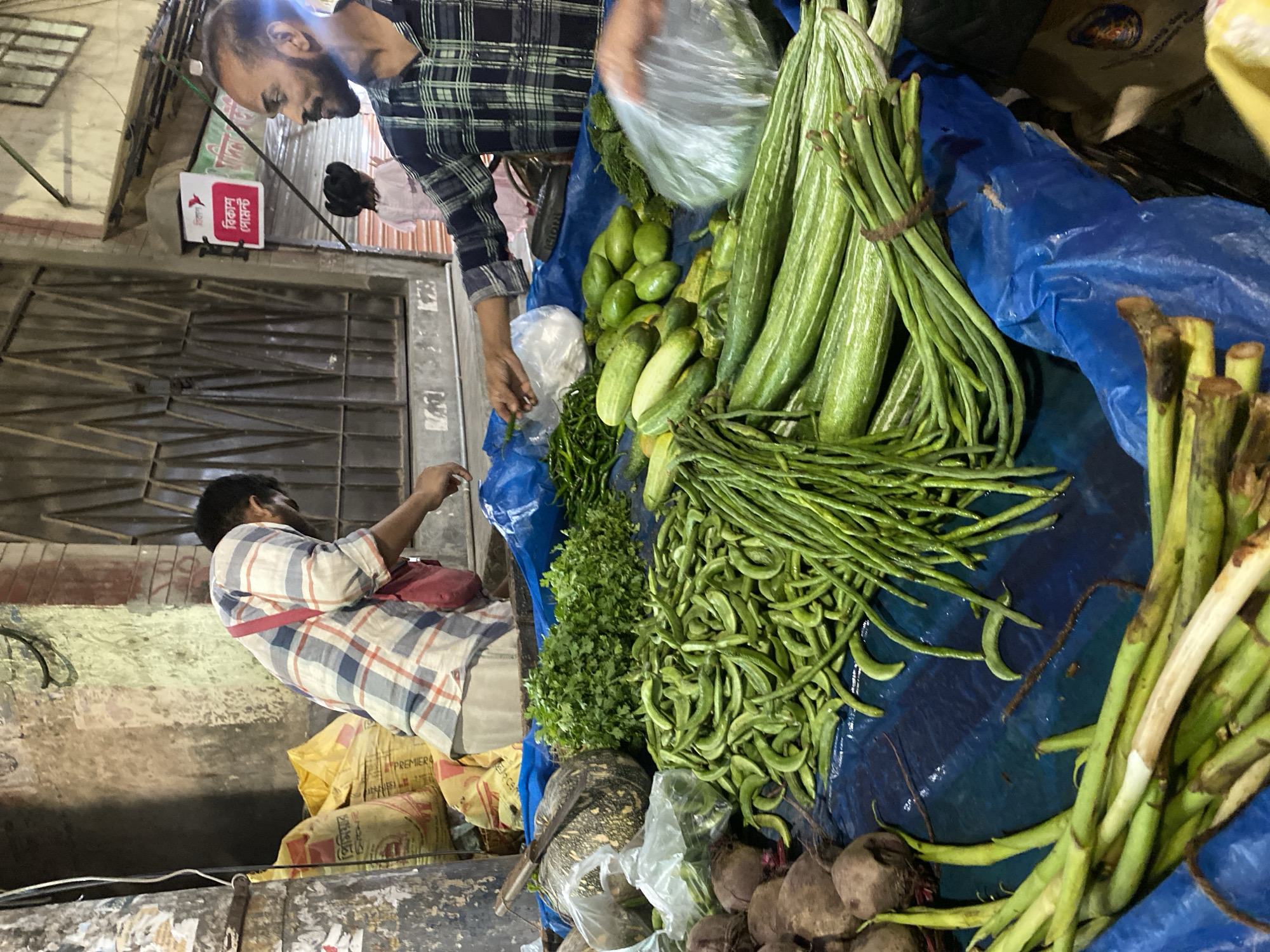 The image depicts a vegetable vendor at a market stall in Dhaka, Bangladesh. The vendor, wearing a plaid shirt, is attending to a customer who is seated and also wearing a plaid shirt. The stall displays a variety of vegetables including snake gourd, beans, cucumbers, other green vegetables, and beets. The vegetables are arranged on a blue tarp. A building is in the background. There are bags of product underneath the seated man.