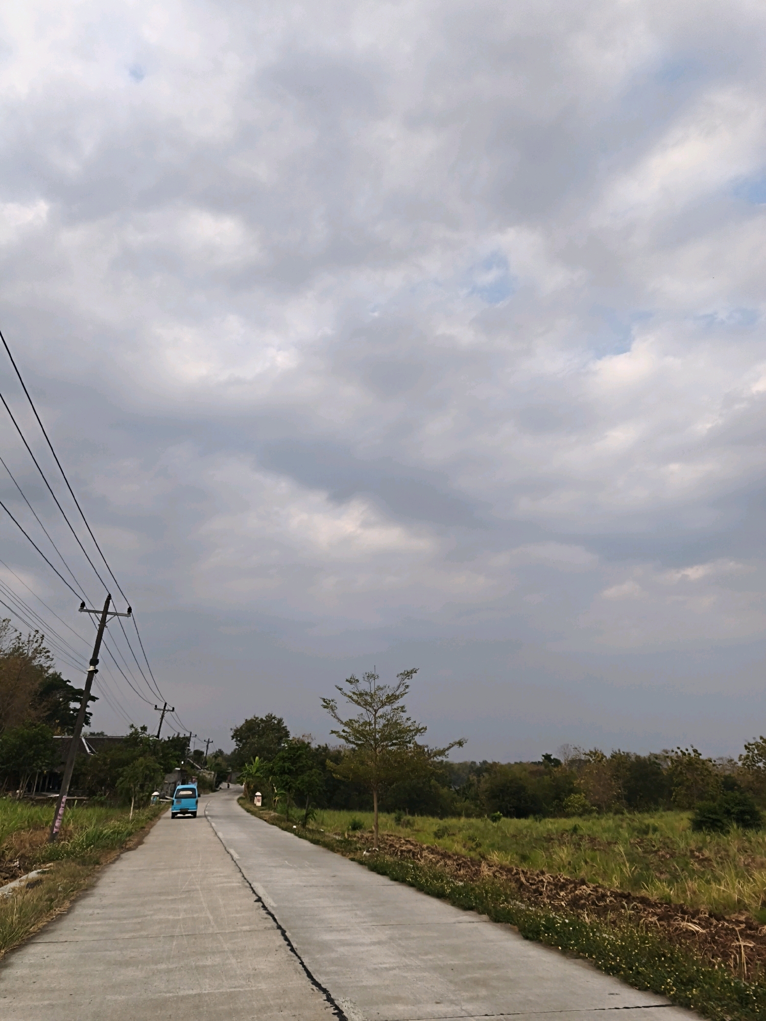 The image depicts a paved road in Krendowahono, Indonesia, under a cloudy sky. On the left side of the road, a utility pole with multiple wires runs parallel to the street. Further down the road, a small blue vehicle is visible. The landscape to the right of the road consists of fields with grasses and some soil. Trees are present on both sides of the road, with denser foliage in the background. The road appears to be in a rural or semi-rural setting, with a few structures visible in the distance. The overall atmosphere is overcast and subdued.