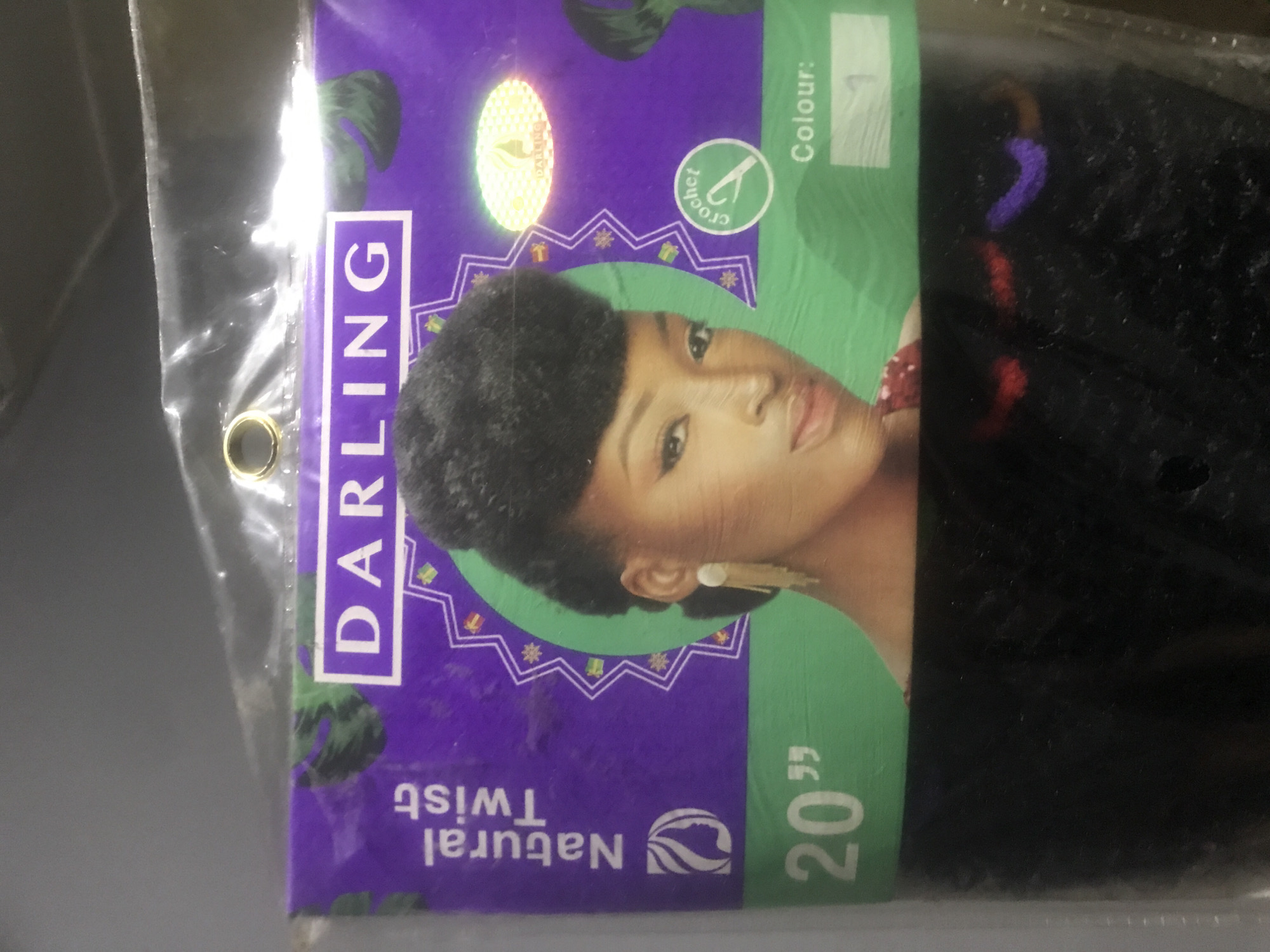 A close-up image, oriented vertically, displays a package of "Darling Natural Twist" hair extensions. The product is encased in clear plastic, which shows some crinkling and glare from the lighting. A brass-colored eyelet is visible at the top left corner of the packaging, suggesting it is designed for hanging.
The packaging features a vibrant label predominantly colored purple and green. The top half of the label is purple, transitioning to a green section on the right side. On the purple background, the brand name "DARLING" is prominently displayed vertically in white capital letters along the left edge. Below it, "Natural Twist" is written horizontally in white. A stylized logo, resembling a wave or 'S' shape, appears next to "Natural Twist".
A striking image of a woman's face, with her head tilted, occupies the center-right of the label, partially overlapping the purple and green sections. She has short, curly dark hair with bangs, light brown skin, and is wearing an earring. Her expression is neutral to slightly pleasant.
Other visible text and symbols on the label include "20''" (likely indicating length in inches) written vertically in white on the green section. Above this, "Colour:" is visible with a blank space next to it, presumably for a color code. A small circular icon with the word "crochet" and a hook symbol is present on the green part. A holographic sticker, which reflects rainbow colors, is visible on the purple section, also bearing the "DARLING" name. Small decorative patterns resembling stars and gift boxes adorn the purple background.
Inside the clear plastic packaging, bundles of synthetic hair are visible. The hair appears predominantly black, but distinct strands of red and purple are interspersed, suggesting a multi-toned product.
The scene is static, focusing entirely on the product packaging, with a plain, dark, out-of-focus background. No specific location cues for Rafin Par, Nigeria, are visually evident in the image itself, though Darling is a prominent brand in the African market.