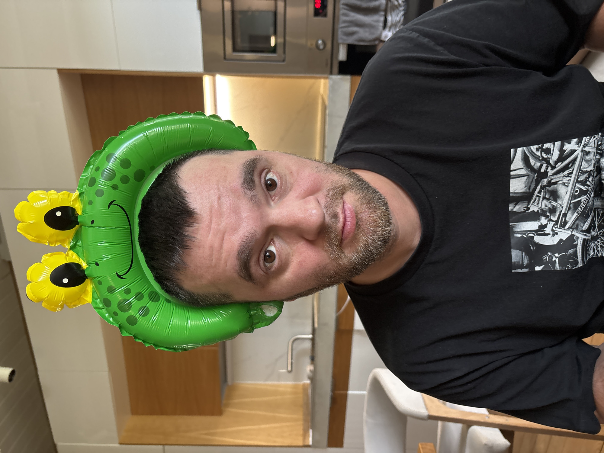 A man with brown hair and a beard wears a green inflatable frog headband with yellow eyes and a smiling mouth. He is looking at the camera with a neutral expression. He is wearing a black t-shirt with a black and white graphic design on the front. The background appears to be a kitchen with wooden cabinets, a countertop with a faucet, and a stainless steel oven. The setting is indoors, likely a residential kitchen. There is no specific indicator of the location being Chaikovsky, Russia.
