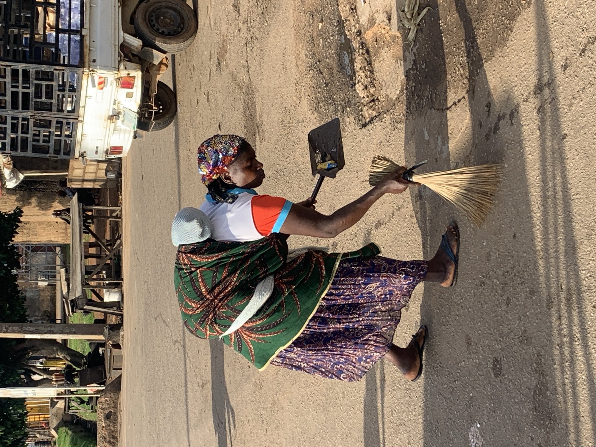 In Gura Top, Nigeria, a woman is sweeping the road, carrying a baby on her back. She is dressed in a vibrant, patterned fabric wrapped around her body, a blue headscarf, and a shirt with orange and blue sleeves. The baby is covered with a light blue cloth. The woman is using a broom made of natural fibers and a dark-colored dustpan. The ground is paved with asphalt, with some debris and cracks visible. In the background, a white truck with a metal cage is parked, and there are buildings, trees, and other people in the distance. The scene is illuminated by natural sunlight, casting shadows on the ground.