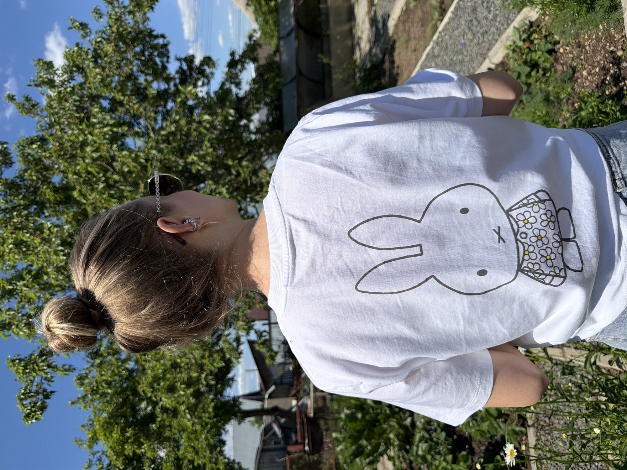 The image displays an individual, viewed from behind, standing outdoors in a garden or yard setting in the city of Chaikovsky, Russia. The person has light brown hair with blonde highlights, styled in a high bun. Their head is turned slightly to the right (viewer's left), revealing the side of their face and ear. Sunglasses with a distinctive silver chain-link design on the arms are worn, resting on the right ear, which also features a silver cuff earring with small dangling elements and a gold-colored stud or hoop.
The individual is wearing a white short-sleeved t-shirt. The back of the t-shirt features a large, black outline illustration of Miffy the bunny, depicted wearing a dress adorned with a white floral pattern and yellow centers. The lower portion of light blue denim jeans is visible at the waistline.
The background features a vibrant green deciduous tree prominently in the middle ground, with smaller green plants and shrubs populating the foreground and mid-ground. A single white daisy-like flower is visible on the left side of the foreground. The ground consists of patches of dark soil, gravel paths, and low-growing vegetation, including what appear to be small red berries, possibly strawberries, on the right. In the distant background, on the left, several rustic or utilitarian buildings with pitched roofs are visible. On the right, further back, structures resembling greenhouses or covered garden beds can be seen. The scene is illuminated by strong, natural sunlight under a clear blue sky with sparse white clouds.