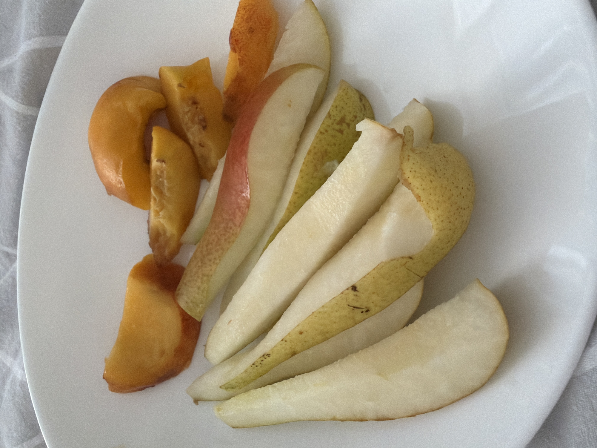A white plate holds slices of fruit on a white textured surface. The fruit consists of pear slices and what appear to be apricot or peach slices. The pear slices are predominantly pale yellow and white, with some exhibiting reddish-pink skin on one side. The apricot or peach slices are a vibrant orange-yellow color. The plate is angled with the top edge curving out of frame. The background is a white, subtly textured textile. There are no people or animals visible in the image. The image does not provide any information about the location of Pazardzhik, Bulgaria.