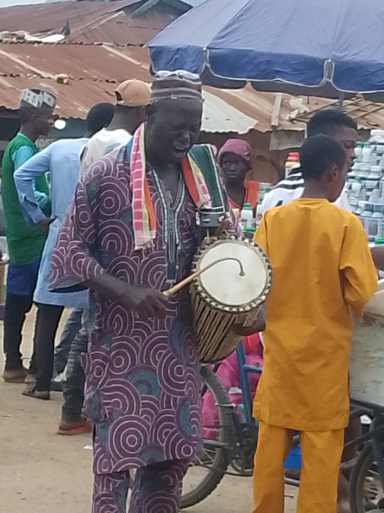 An outdoor market scene in Komu, Nigeria, features a man playing a djembe drum. He is wearing a vibrantly patterned purple and white traditional outfit and a colorful scarf around his neck. To his right, a young person in an orange robe stands facing away from the camera. Several other individuals are visible in the background, some wearing traditional attire and hats. The market appears to be set up under a blue awning, with corrugated metal roofs and stalls visible. The ground is unpaved, suggesting a dusty or sandy surface. A bicycle is parked to the left of the drummer. The overall impression is of a lively community gathering or event.