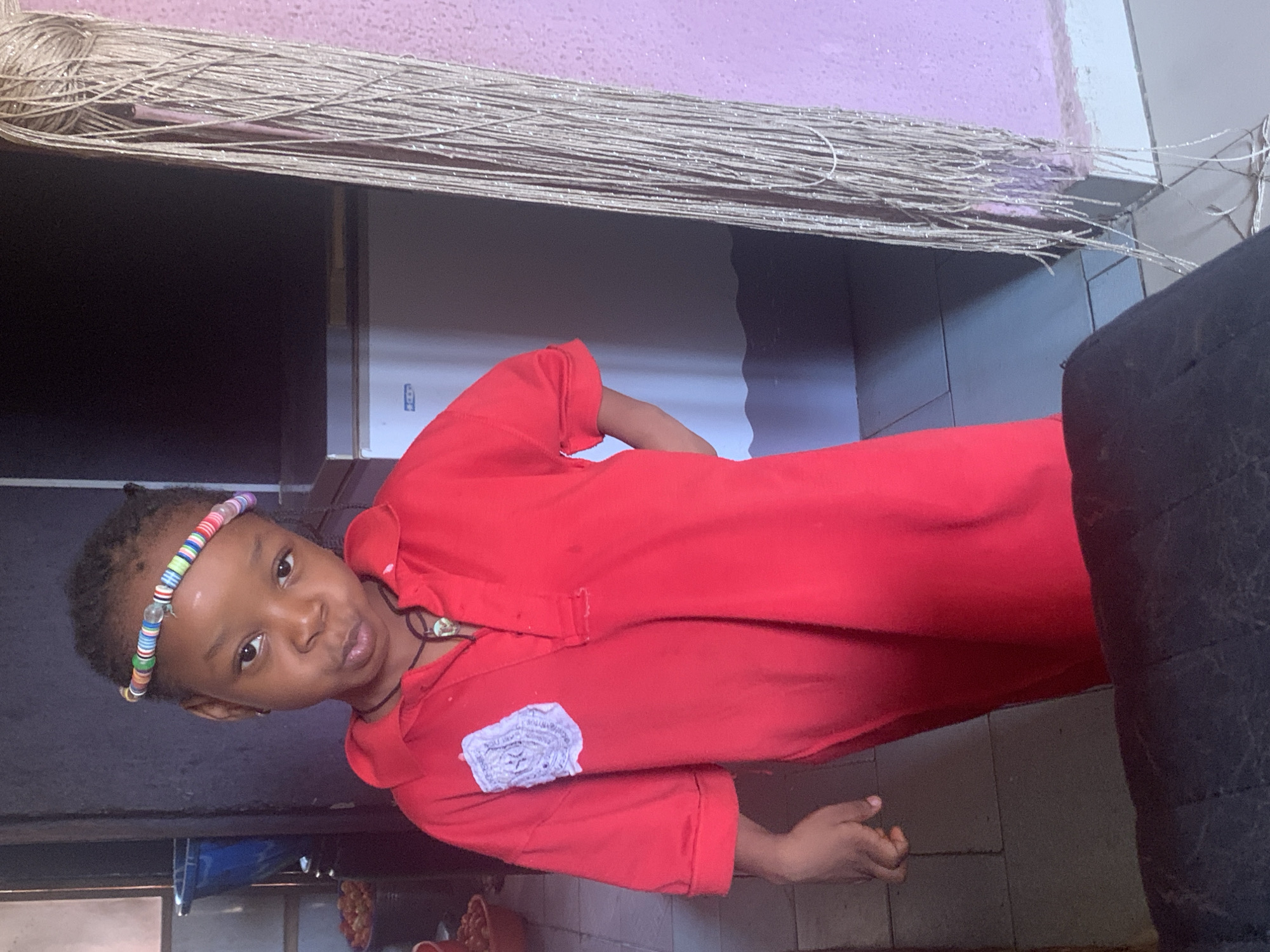 A young, dark-skinned child is shown in the foreground of the image. They are wearing a bright red long-sleeved shirt or dress with a beaded headband and a necklace. A light-colored, possibly silver string-like decoration hangs down from above. Behind the child are a refrigerator and walls painted in shades of pink and white/grey. The flooring appears to be tiled. A portion of a person seated or lying down wearing black clothes is visible on the right-hand side. The location is in Sabon Tasha, Nigeria.