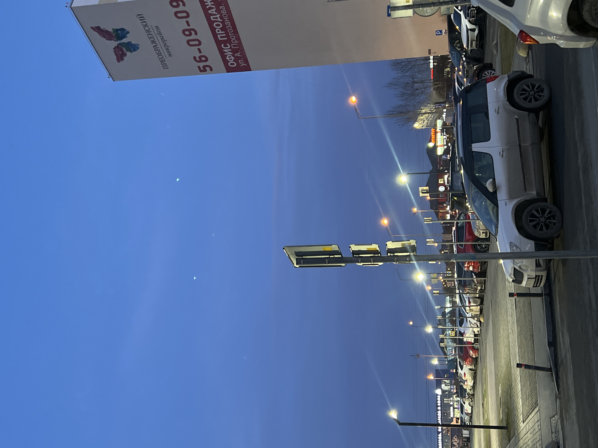 The image shows a street in the city of Tyumen, Russia, at dusk. On the right side of the image is a street lined with parked cars. Several lampposts line the street, casting light onto the road and cars. In the background, there are buildings and signs for shops, including "MORETO". On the left is a large banner with the text "Преображенский микрорайон" (Preobrazhensky microdistrict) and a phone number "56-09-09" and "ОФИС ПРОДАЖ ул. А. Протозанова." (Sales office, st. A. Protozanova). The sky is a gradient of deep blue near the top to a lighter orange near the horizon. A traffic light is visible in the middle of the image.