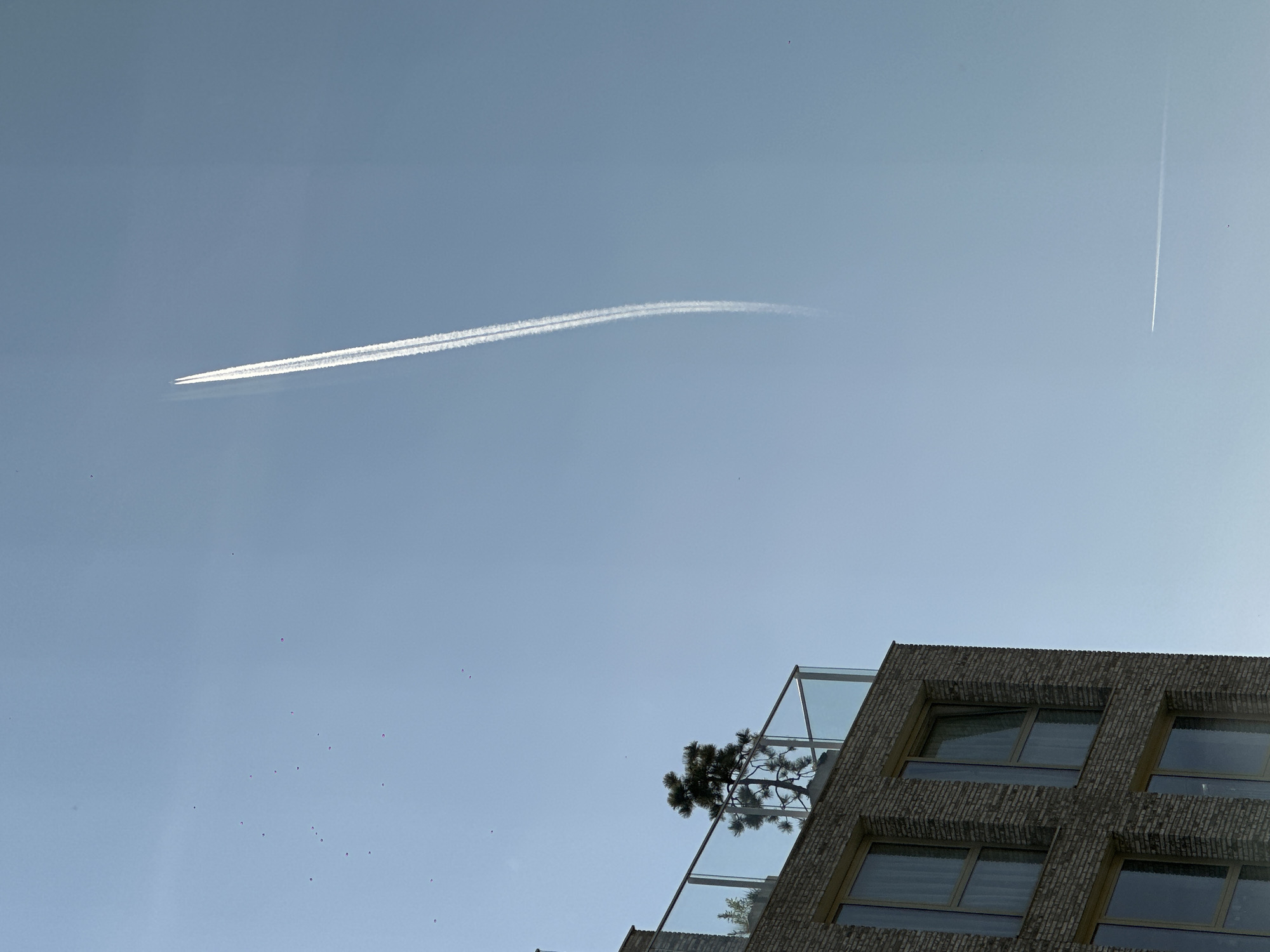 The image captures a clear, blue sky in Amsterdam, Netherlands. Two contrails are visible, one extending horizontally across the sky and the other oriented vertically. In the lower right of the frame, the top of a modern building is visible. It features a brick facade, rectangular windows, and a balcony with a glass railing and trees. Several small dark spots, possibly birds or insects, are scattered across the sky.