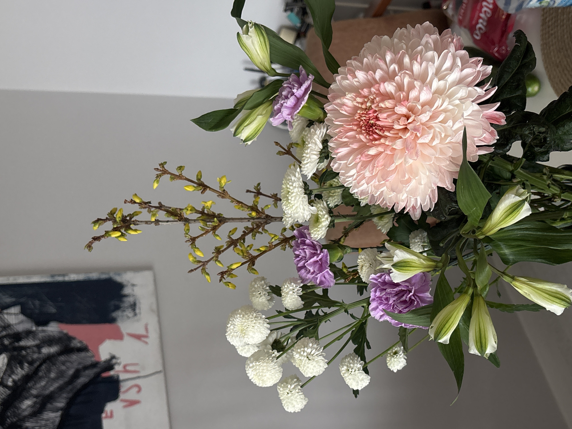 The image shows a bouquet of flowers. Dominating the bouquet is a large, pale pink chrysanthemum-like flower. There are also purple carnations, small white round flowers, and unopened lily buds with green leaves. Thin branches with small yellow buds extend from the bouquet. The bouquet is positioned in front of a painting with black, white, and red colors, and a gray wall is visible in the background. The location of the image is not mentioned. The city of Krasnoyarsk is not mentioned.