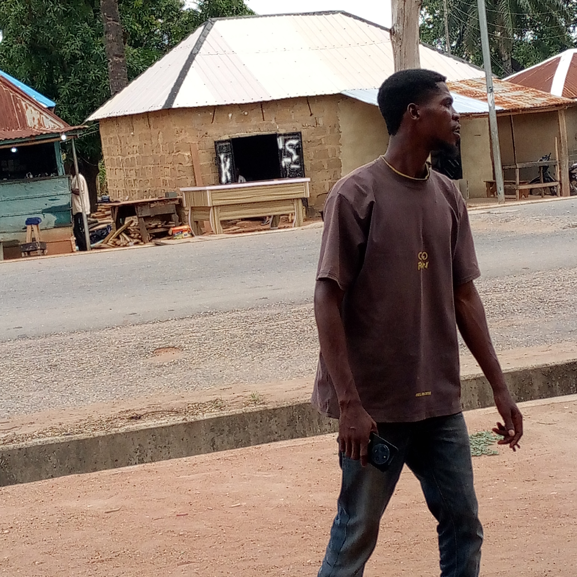 A man is depicted walking or standing on a dirt path next to a paved road, looking towards the right side of the frame. He has dark skin, short dark hair, and a slight beard, and wears a dark brown short-sleeved t-shirt with a gold-colored design on the left chest that reads "CO RH." A smaller, less distinct word, possibly "GLORY," is visible near the bottom hem. He also wears blue jeans and a thin gold-colored chain around his neck. In his right hand, he holds a black smartphone with a multi-lens camera module.
The scene appears to be a street in a rural or semi-urban area. Behind the man, a gray, gravelly road separates him from several buildings. On the left side of the background, a light blue building with a rusty, light blue corrugated metal roof is visible. A person in a light-colored garment stands near it. In the center-back, a building constructed of unrendered concrete blocks or mud bricks features a light-colored corrugated metal roof. Its window opening has dark shutters, with a white letter "K" painted on the left shutter and a stylized "S" or "2" on the right. Below this window, a light-colored wooden structure, possibly furniture or a display, is present. Further to the right, another building with a rusty corrugated metal roof is partially visible. Green foliage from trees dots the background, suggesting a natural environment alongside the structures. The lighting indicates it is daytime.