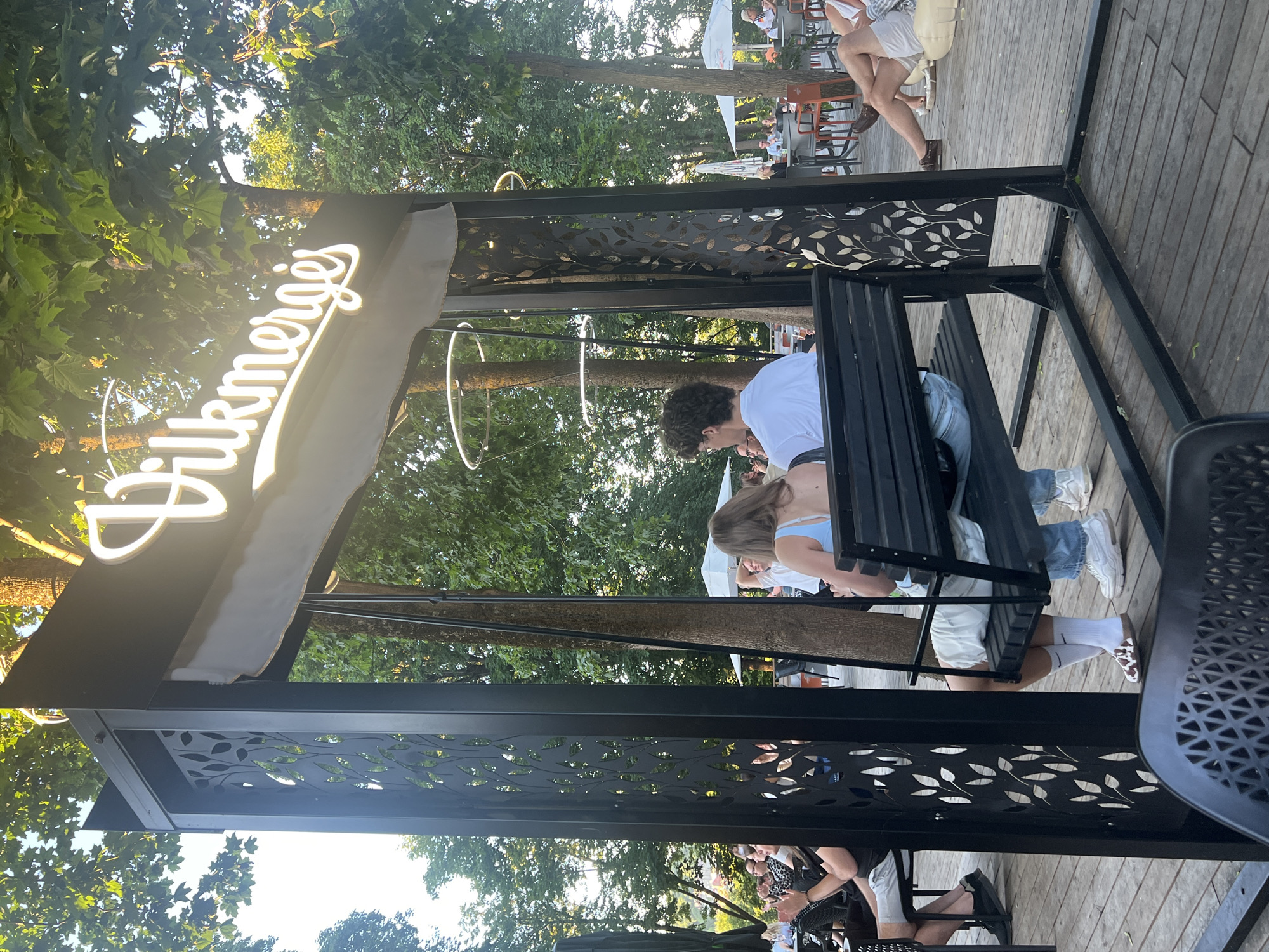 The image shows a swing structure with a neon sign that reads "Vilkmērģis" at an outdoor seating area. Two people, a man and a woman, are seated on the swing, facing away from the camera. The woman is wearing a light blue top and white shorts, while the man is wearing a white t-shirt and glasses. The swing structure is made of black metal with decorative leaf cutouts. In the background, other people are seated at tables and on loungers, with trees and white umbrellas visible. The setting appears to be a park or an outdoor restaurant. The sign indicates the location is likely in Vilnius, Lithuania, as the name "Vilkmērģis" is associated with a restaurant there. The overall scene depicts a relaxed atmosphere, with people enjoying themselves in an outdoor environment.