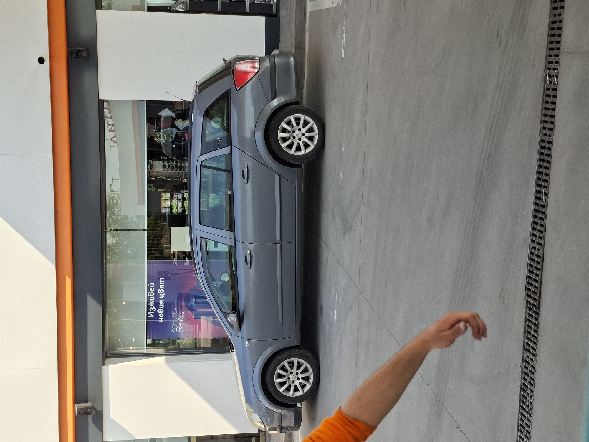 A gray Opel Astra station wagon is parked perpendicularly against the storefront of a commercial building with large glass windows in Plovdiv, Bulgaria. The building has an orange vertical stripe on its facade. A person's arm wearing an orange sleeve is visible in the lower right corner of the frame. A grey concrete pavement stretches from the car to the right side of the image, where a drainage grate runs parallel to the pavement edge. Reflections are visible in the building's windows. An advertising sign inside the building reads "Изживей новия цвят", translated roughly to "Experience the new color".